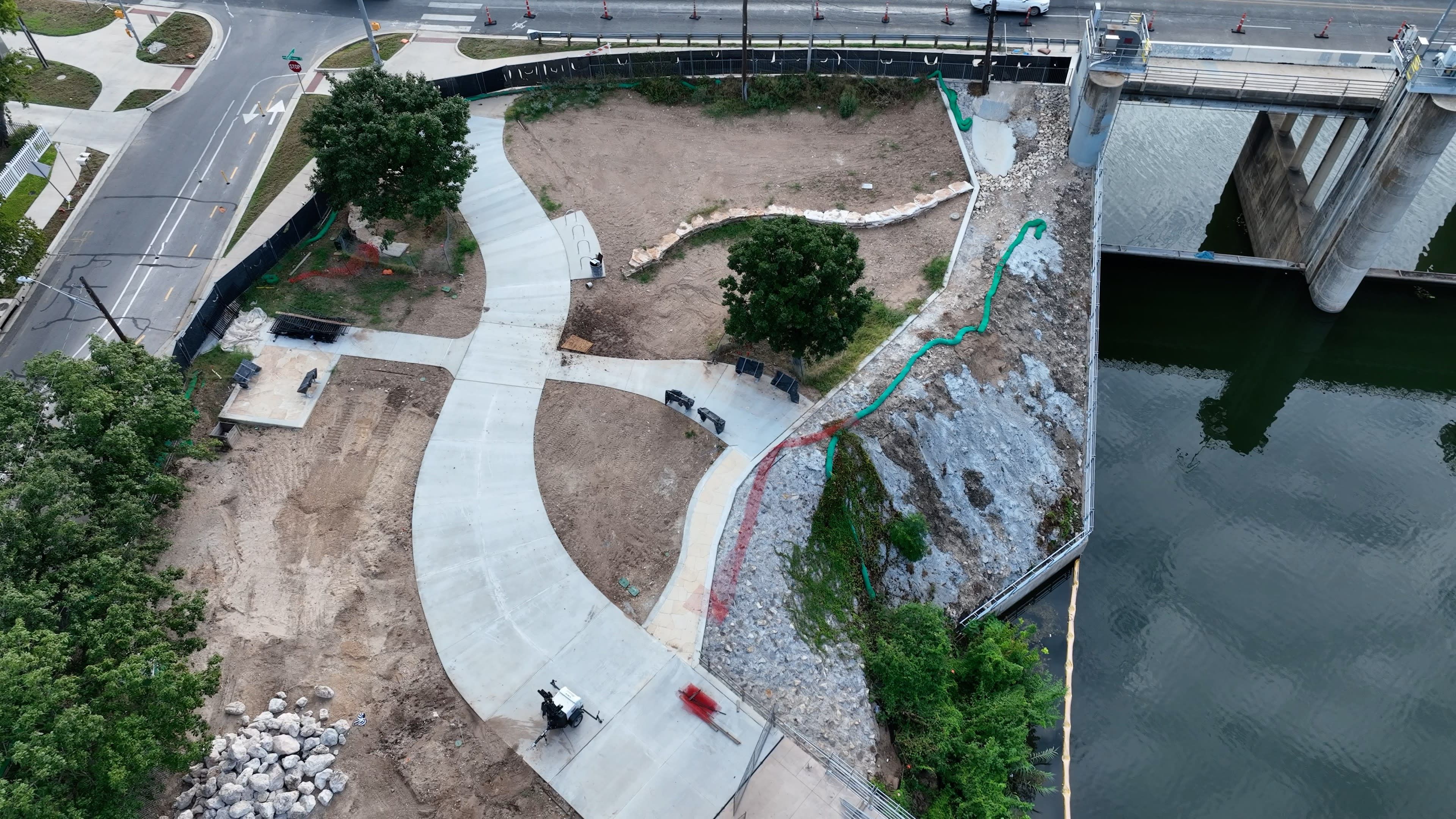 Aerial view of a waterfront park under construction with concrete pathways, scattered trees, benches, rocky slope, and a dam structure over water to the right.