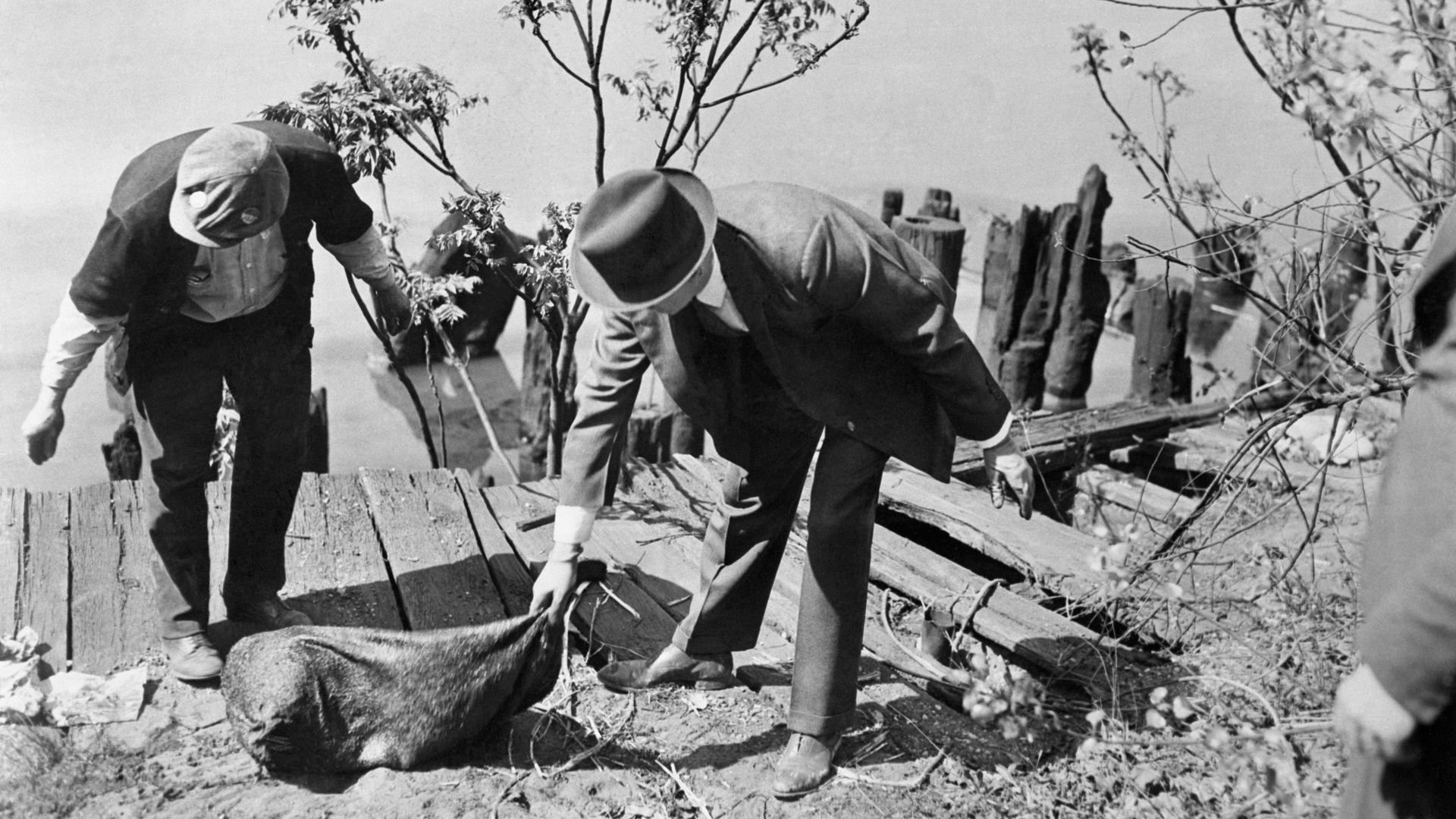 Two men near a wooden dock by water, one in a suit and hat, the other in work clothes, lifting a large sack on a dirt and grass bank with small trees nearby.