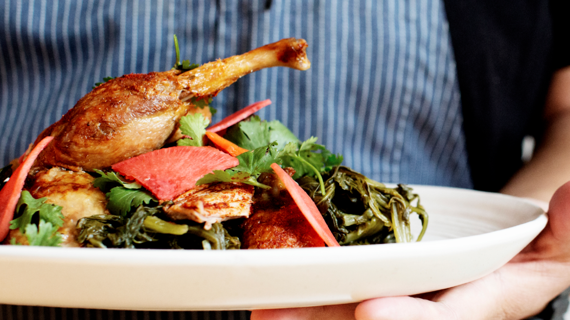 A person holding a plate of food with greens and a cooked chicken leg on top.