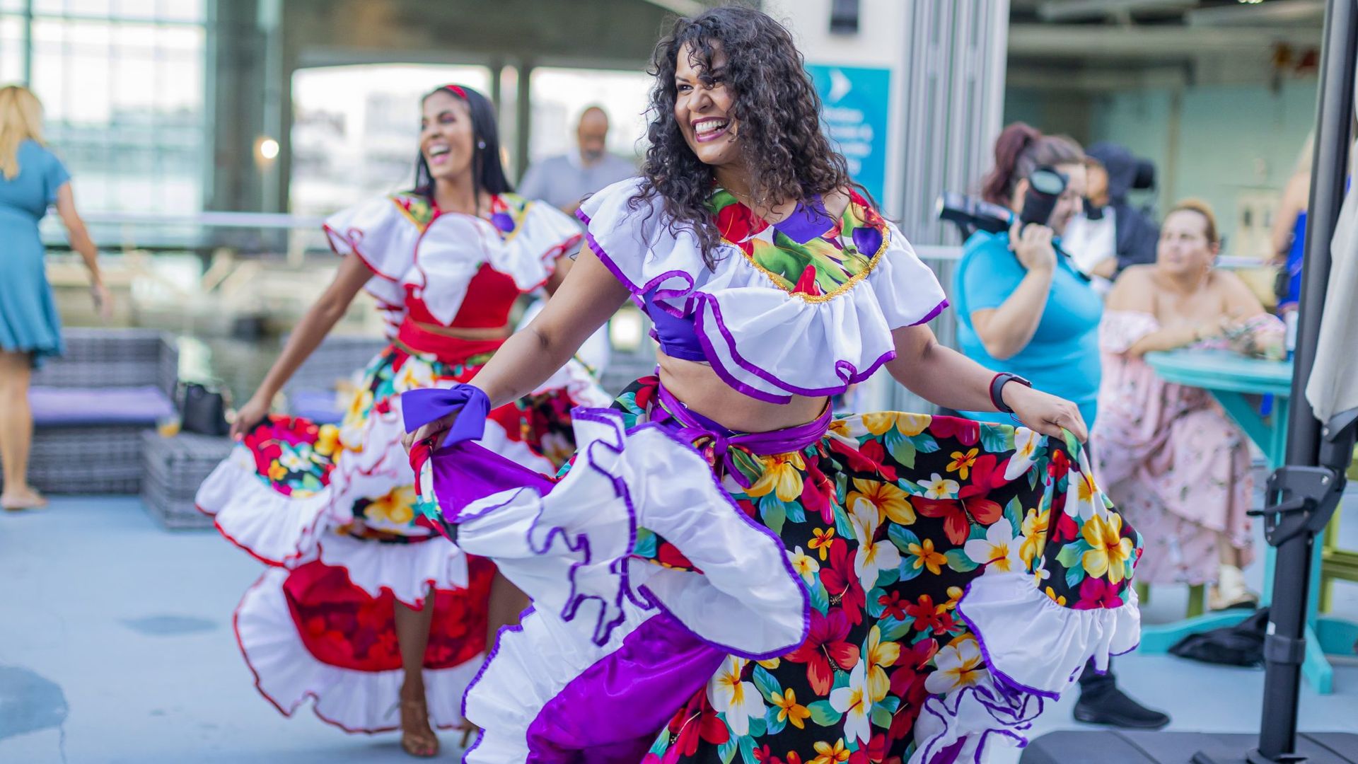 Dancers in bright floral costumes perform on a sunny outdoor deck. Foreground, a smiling woman with curly hair wears a white and purple ruffled top and a vibrant floral skirt.