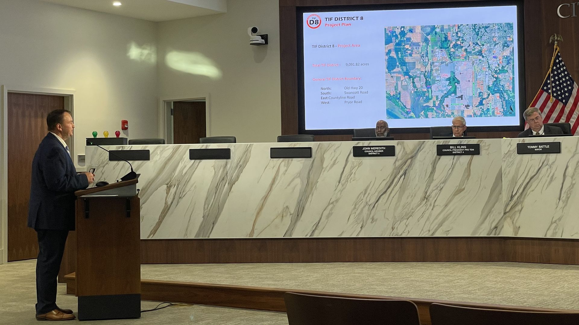 Man in a suit speaks at a podium in a council chamber; a marble desk with nameplates faces a large screen displaying a map and the title "TIF District 8 Project Plan" with an American flag nearby.
