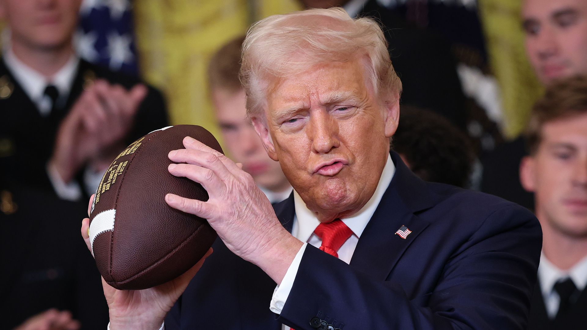 President Donald Trump in blue suit and red tie holds football and purses lips.