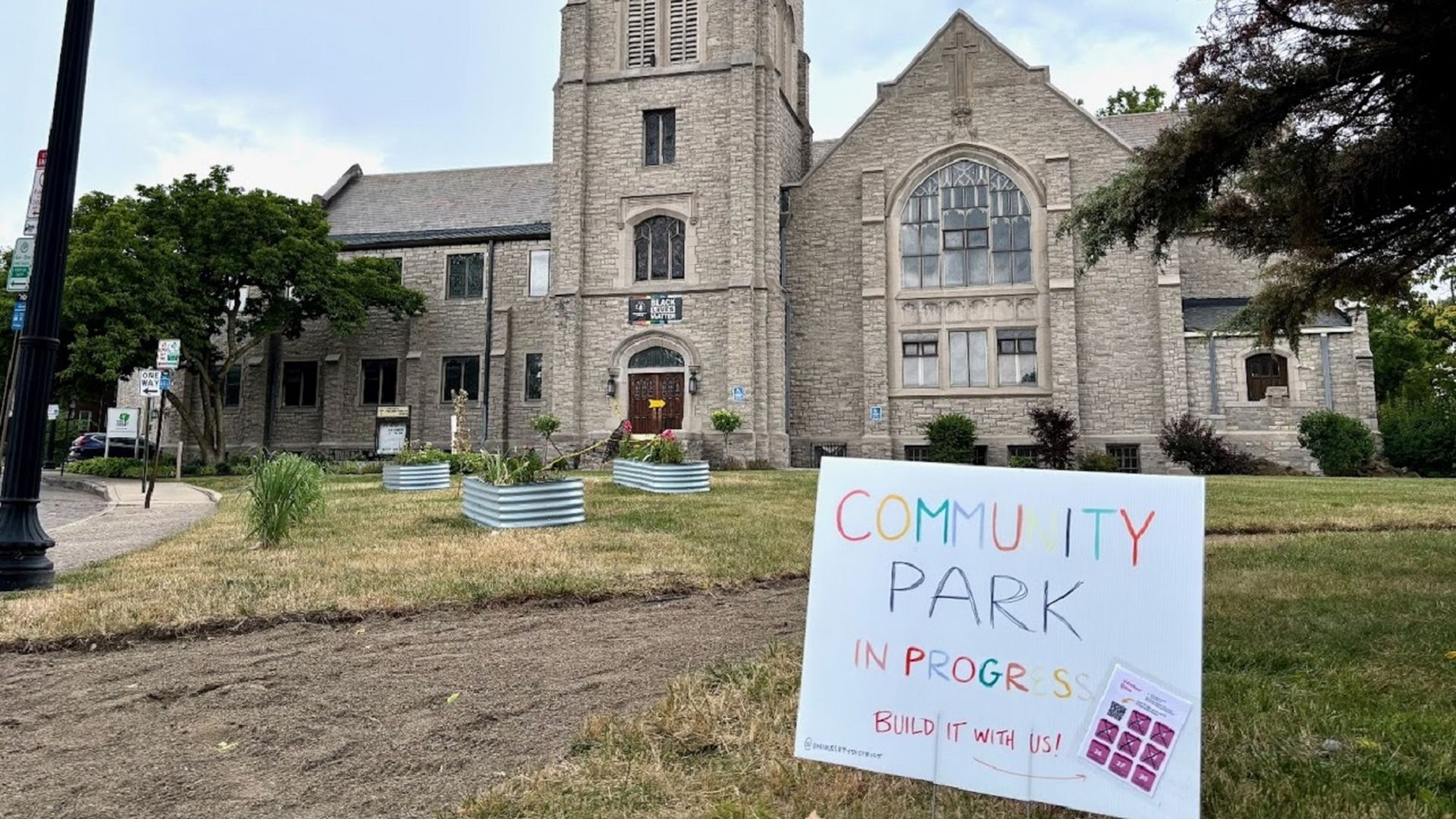 A sign reads "Community Park in Progress, Build It With Us!" in front of a green space on the lawn of a church. 