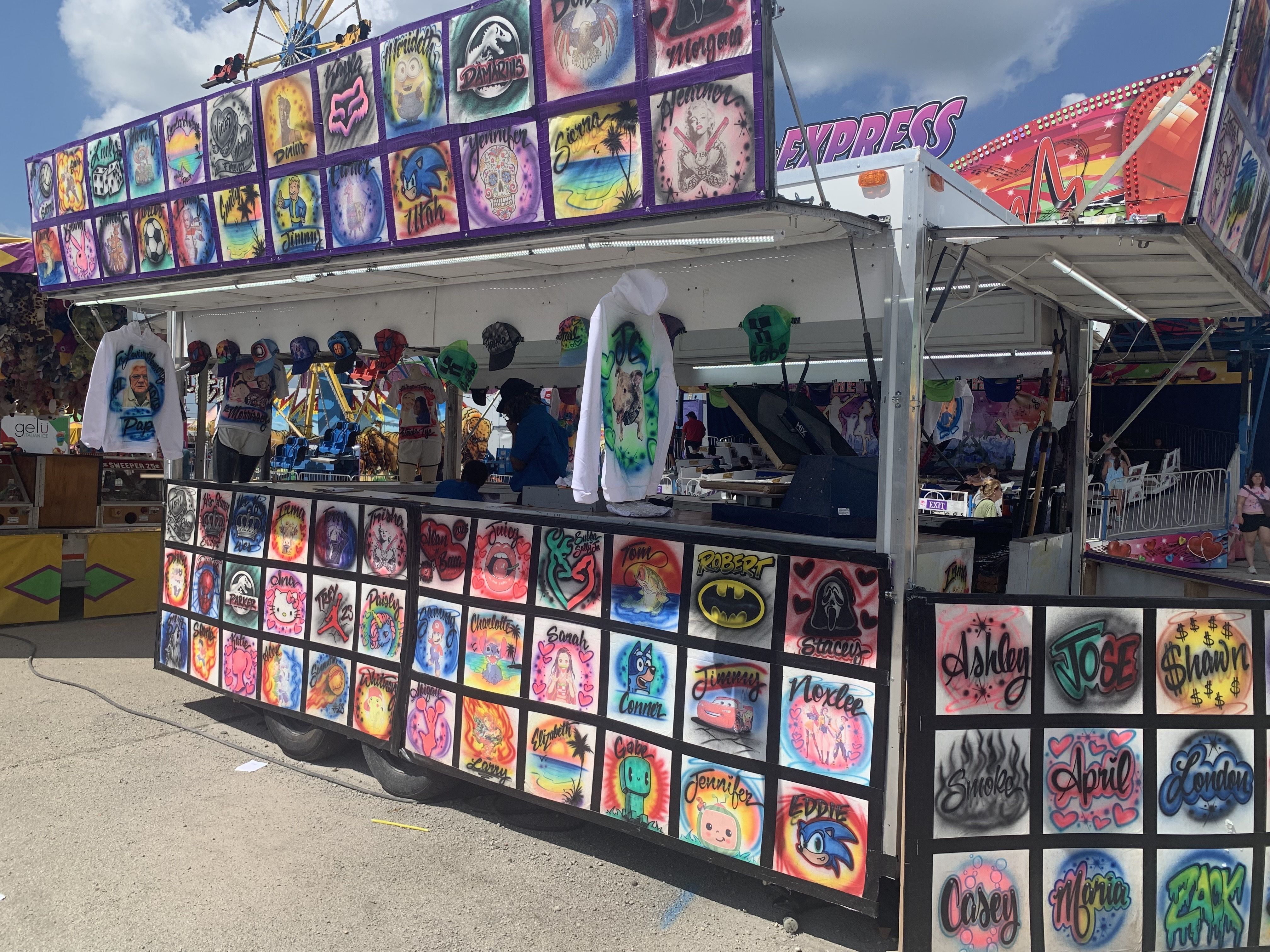 Colorful carnival booth with a wall of square panels featuring cartoon portraits and names, purple framing, bright EXPRESS sign on the right, hats and merchandise visible, people browsing, blue sky overhead.