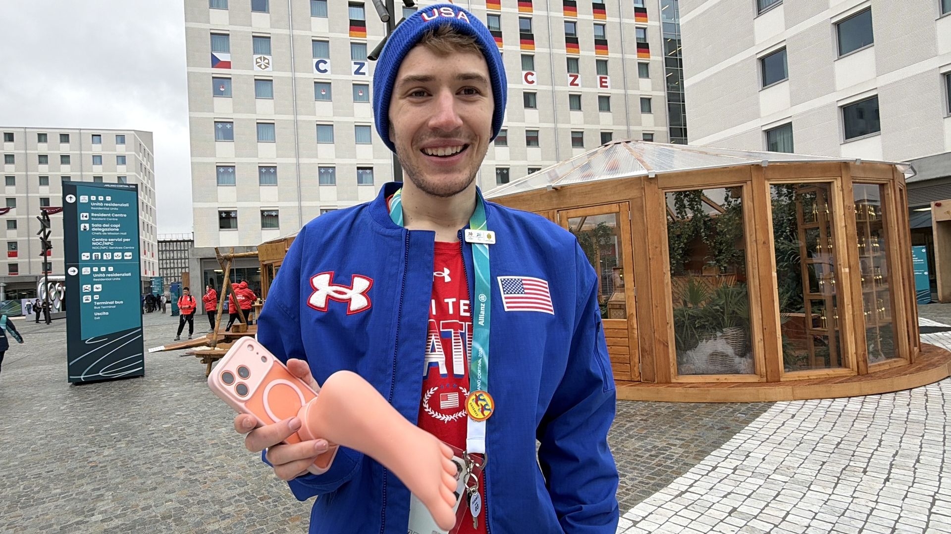 Smiling man in blue USA jacket and hat holds a phone with a pink foot-shaped case, standing outside near modern buildings with colorful window decorations and a wooden gazebo.