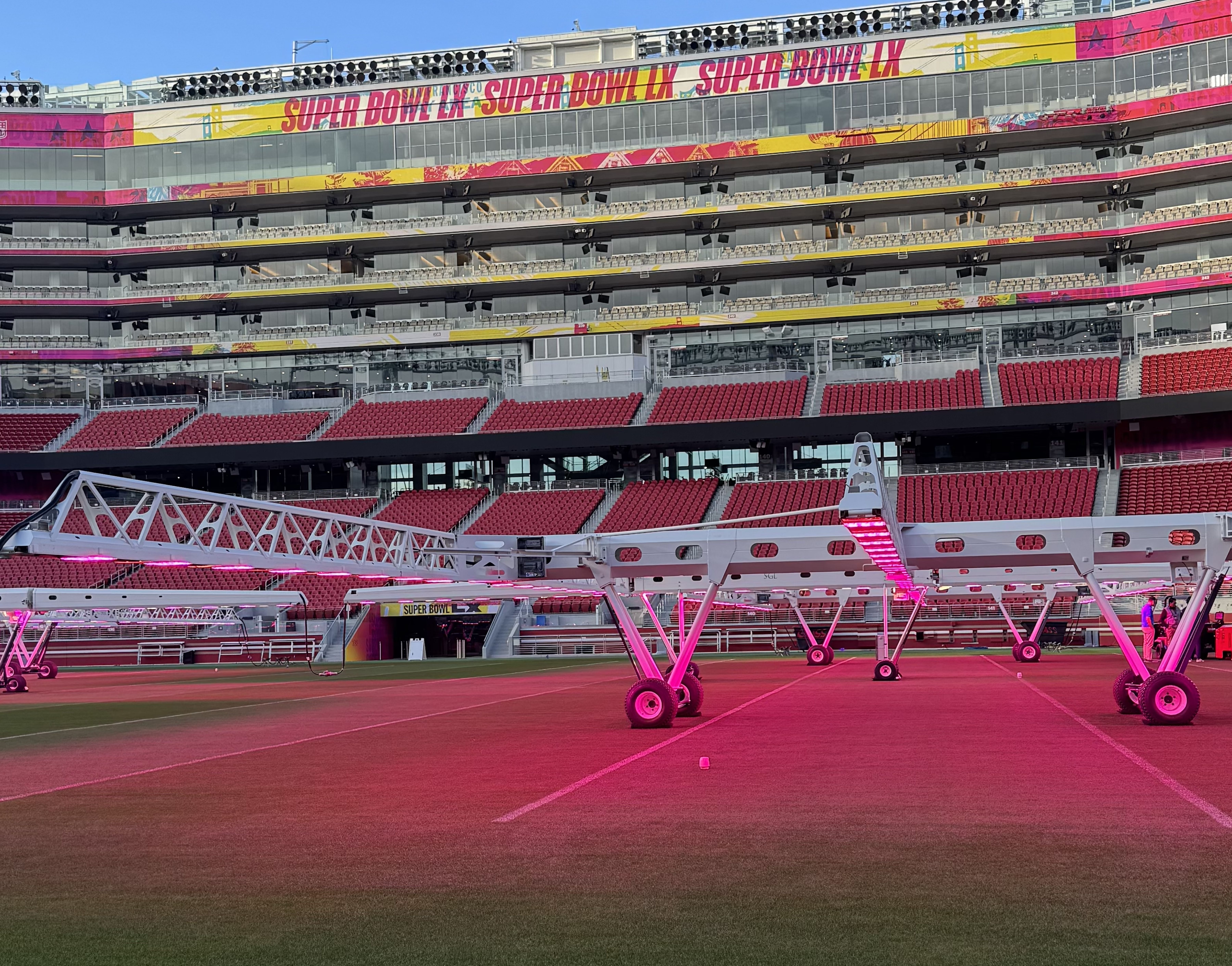 Empty San Francisco stadium with red seats and Super Bowl LX banners, featuring white frames with pink lights on the green football field.