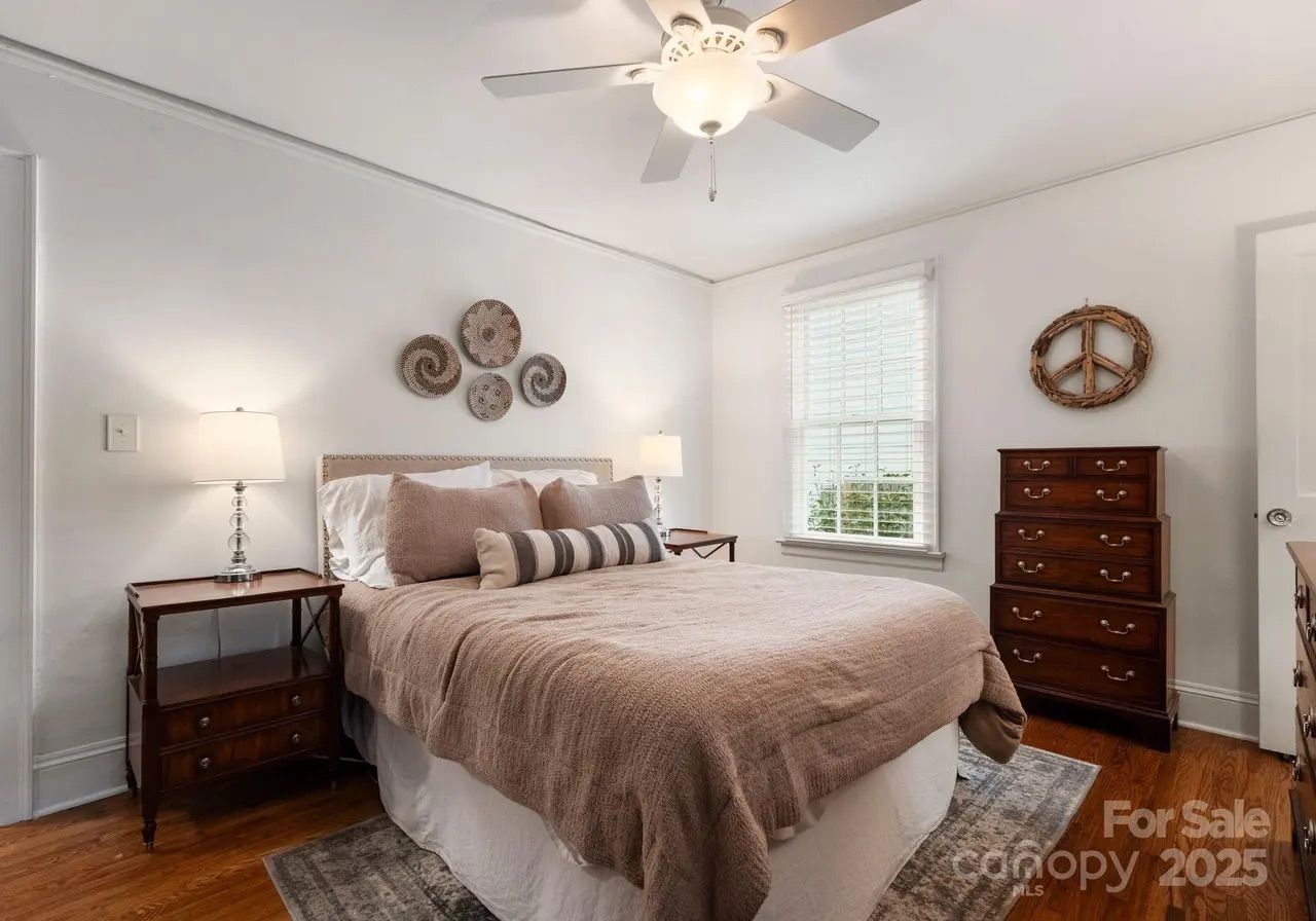 Cozy bedroom with beige bedding, wooden nightstands with lamps, a hardwood floor, a ceiling fan, wall decor including woven plates and a peace sign, and a window with white blinds.