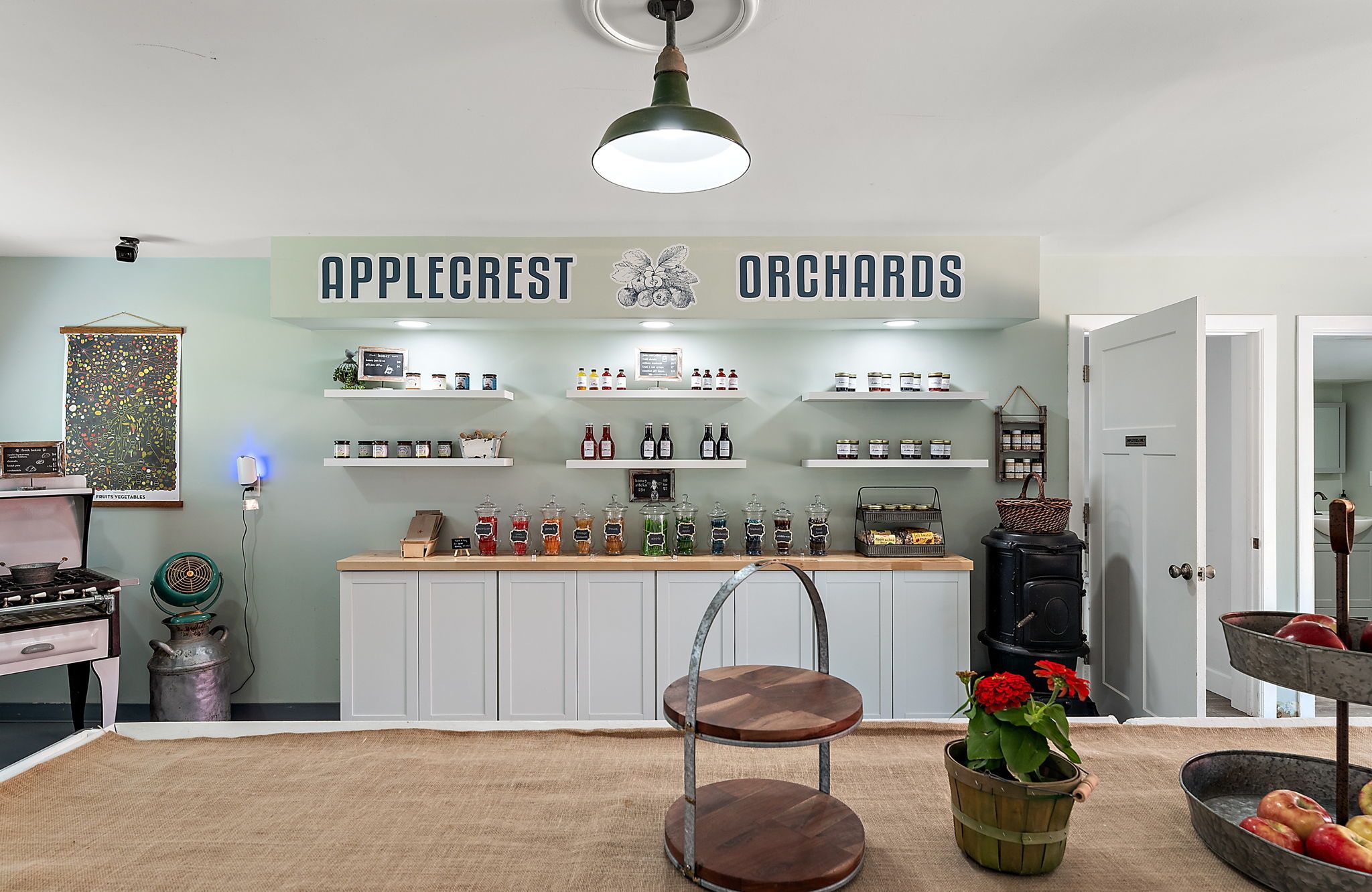 Interior of Applecrest Orchards shop with white cabinets, jars of preserves, sauces on shelves, vintage stove, green wall, hanging light, and red flowers in a small green pot.