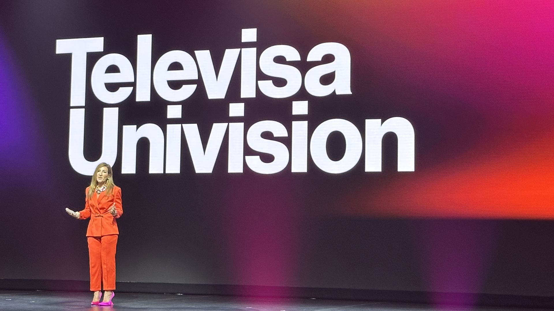 TelevisaUnivision's Donna Speciale in an orange pantsuit with pink high heels speaking in front of a screen that says "TelevisaUnivision"