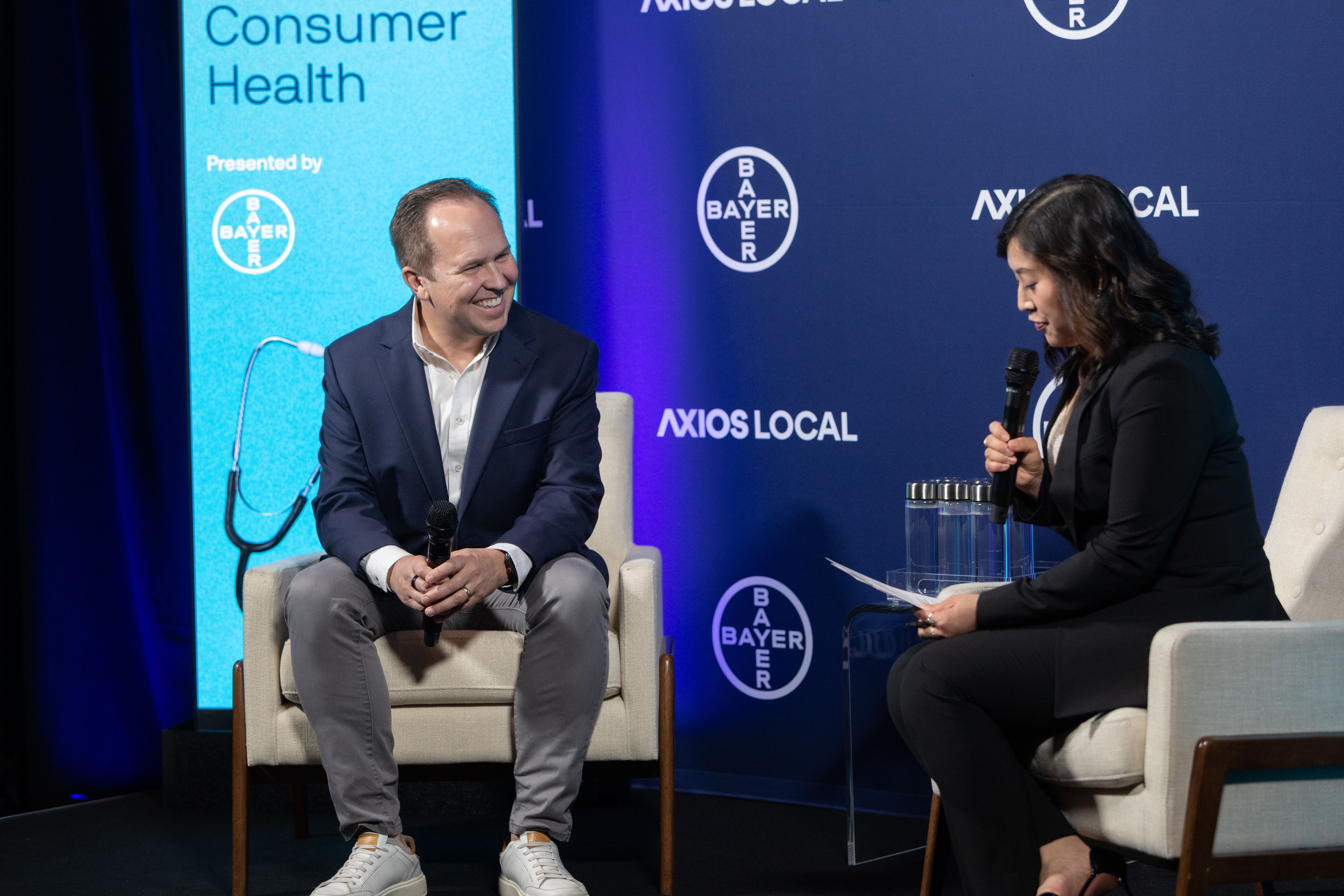 Two panelists on stage with a blue Bayer and Axios Local backdrop; a smiling man in a navy blazer sits left, a woman holds a microphone.