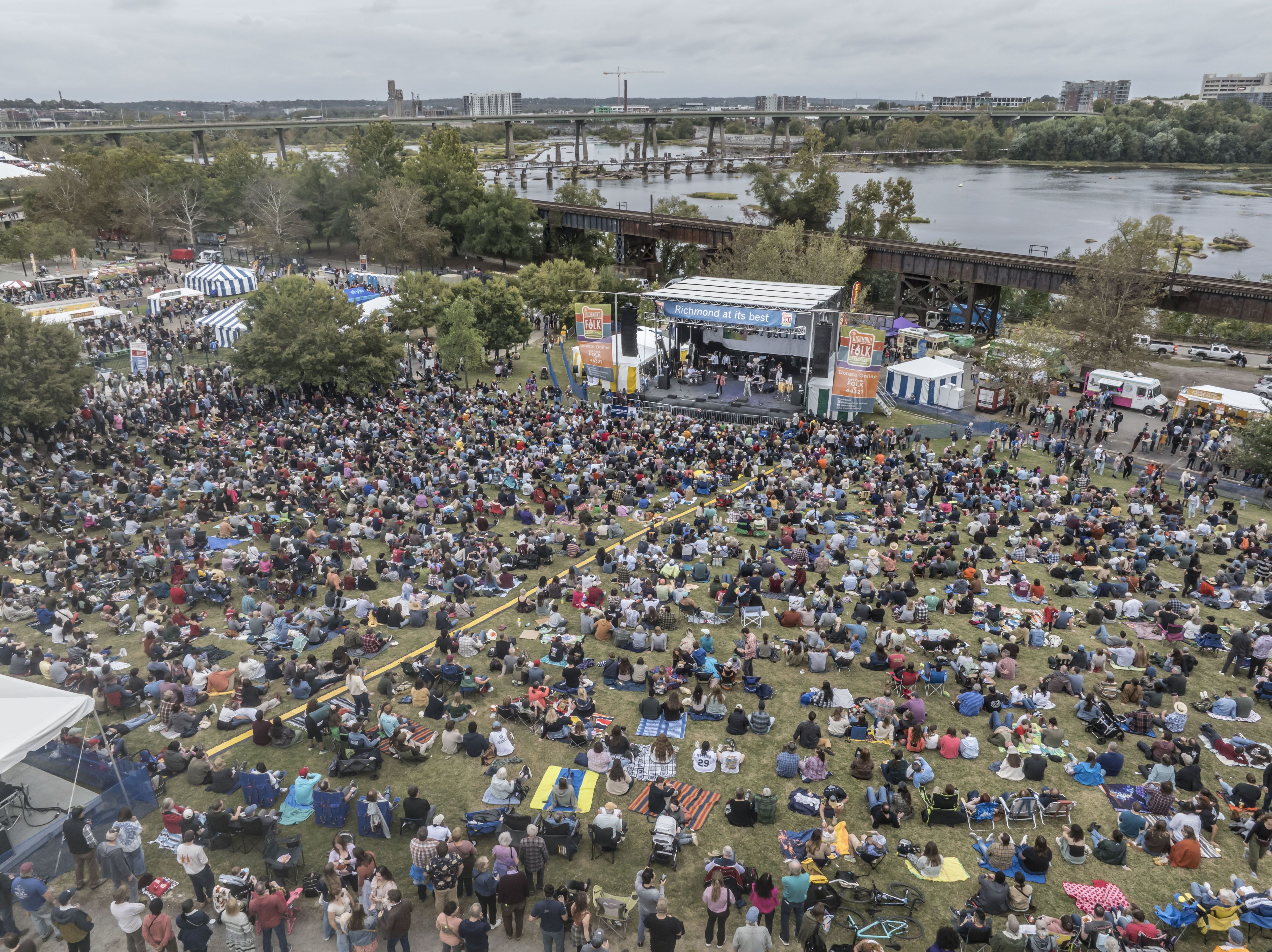 Crowd at an outdoor riverfront concert in a park, blankets on the grass facing a stage that reads "Richmond at its best." Food tents, trees, and a distant bridge in the background.