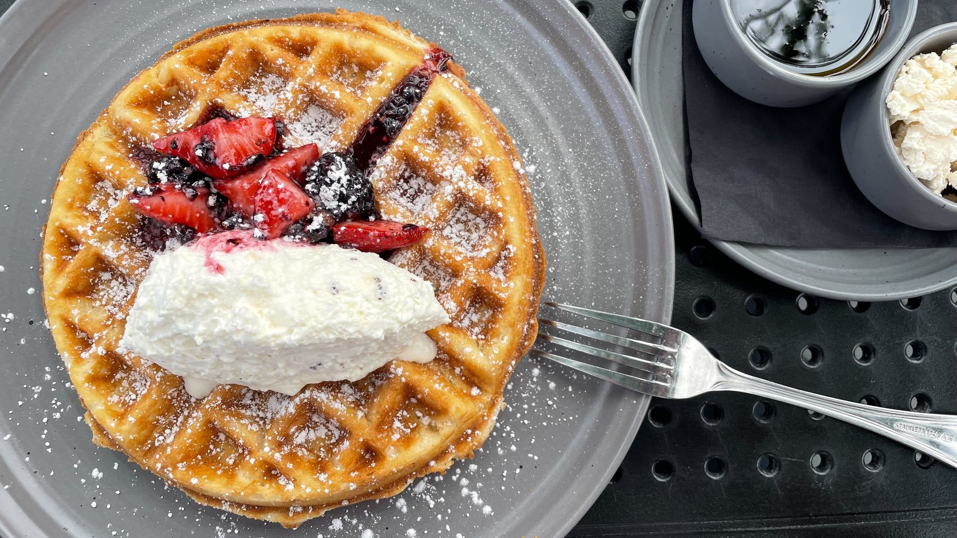 A waffle topped with whipped cream, strawberries and berries, on a gray plate. A fork is nearby.