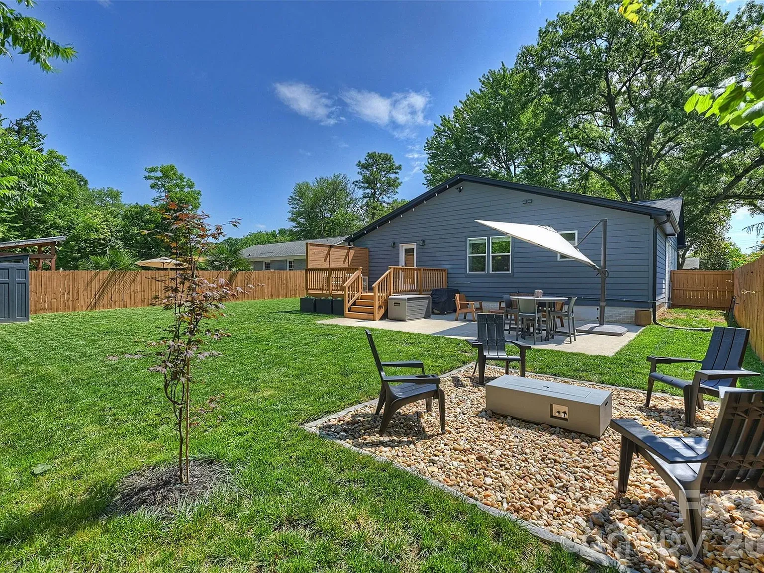 Backyard of a blue house with a wooden deck and patio table under a large umbrella. A gravel fire pit with four chairs sits near the seating area, surrounded by green lawn and tall trees.