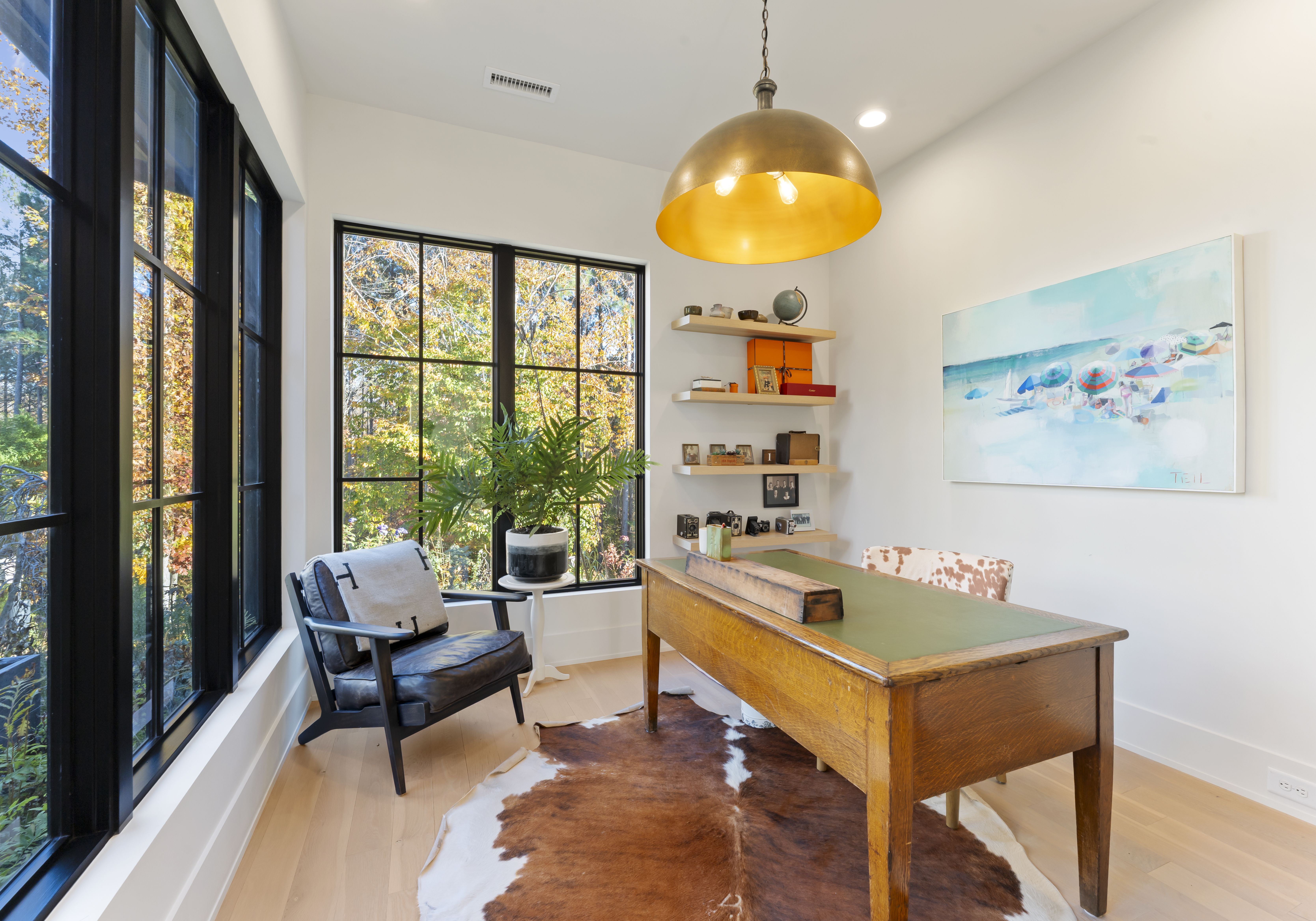 Bright home office with black-framed windows showing autumn trees. Wooden desk with green pad on a cowhide rug; black chair, plant on white side table. Art, floating shelves, and a gold lamp overhead.