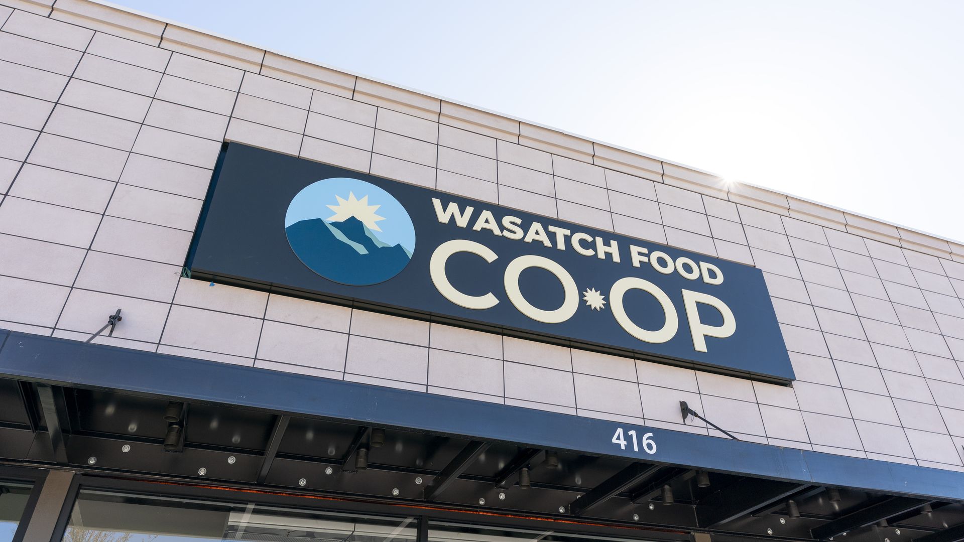 Angle view of a beige tiled storefront with a dark blue sign reading "WASATCH FOOD COOP". A round logo shows sun over mountains; sun glare to the right; 416 on a blue trim.