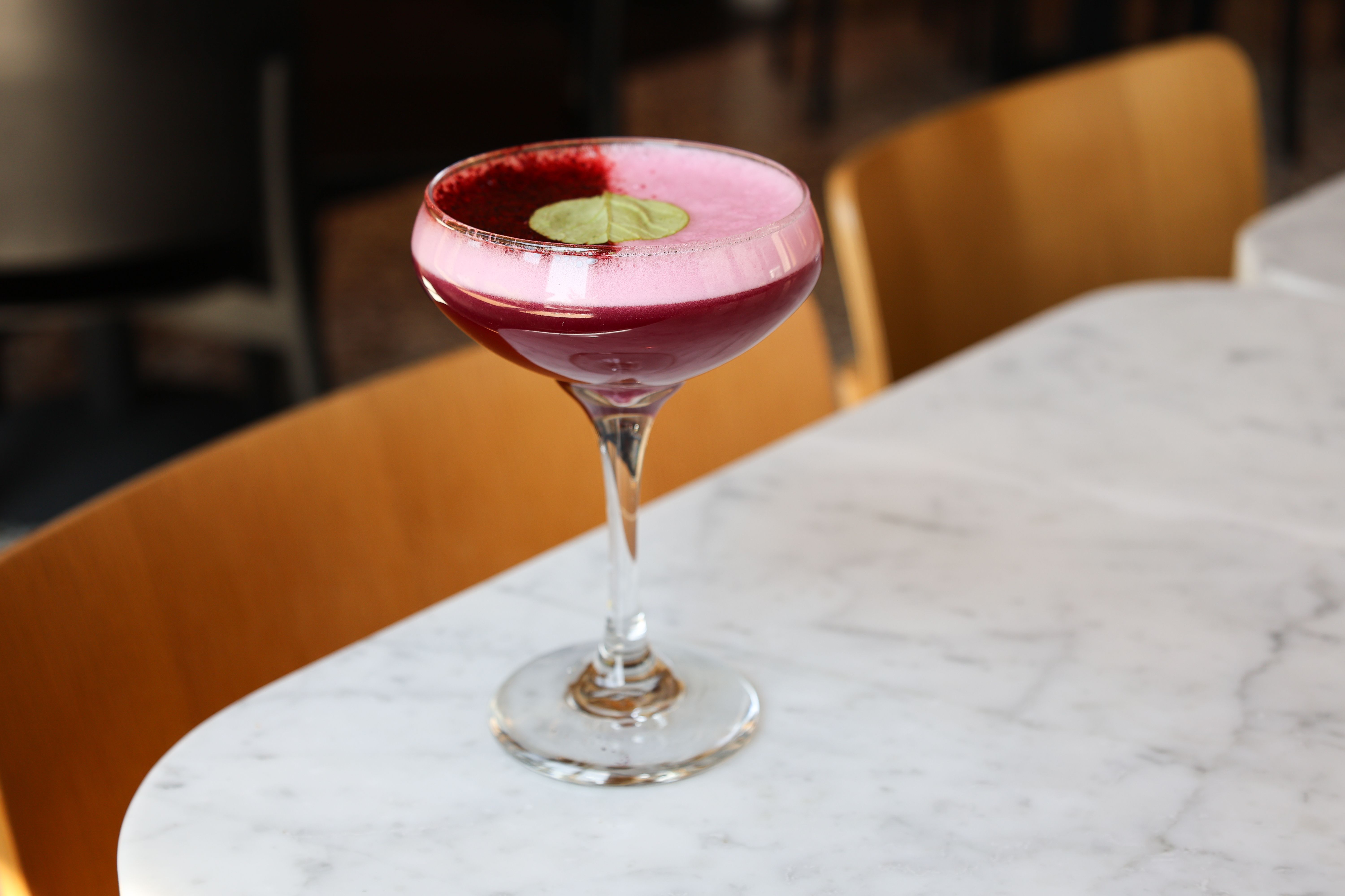 Purple-red cocktail in a coupe glass with pink foam and a green leaf garnish, sitting on a white marble table with wooden chairs in the background