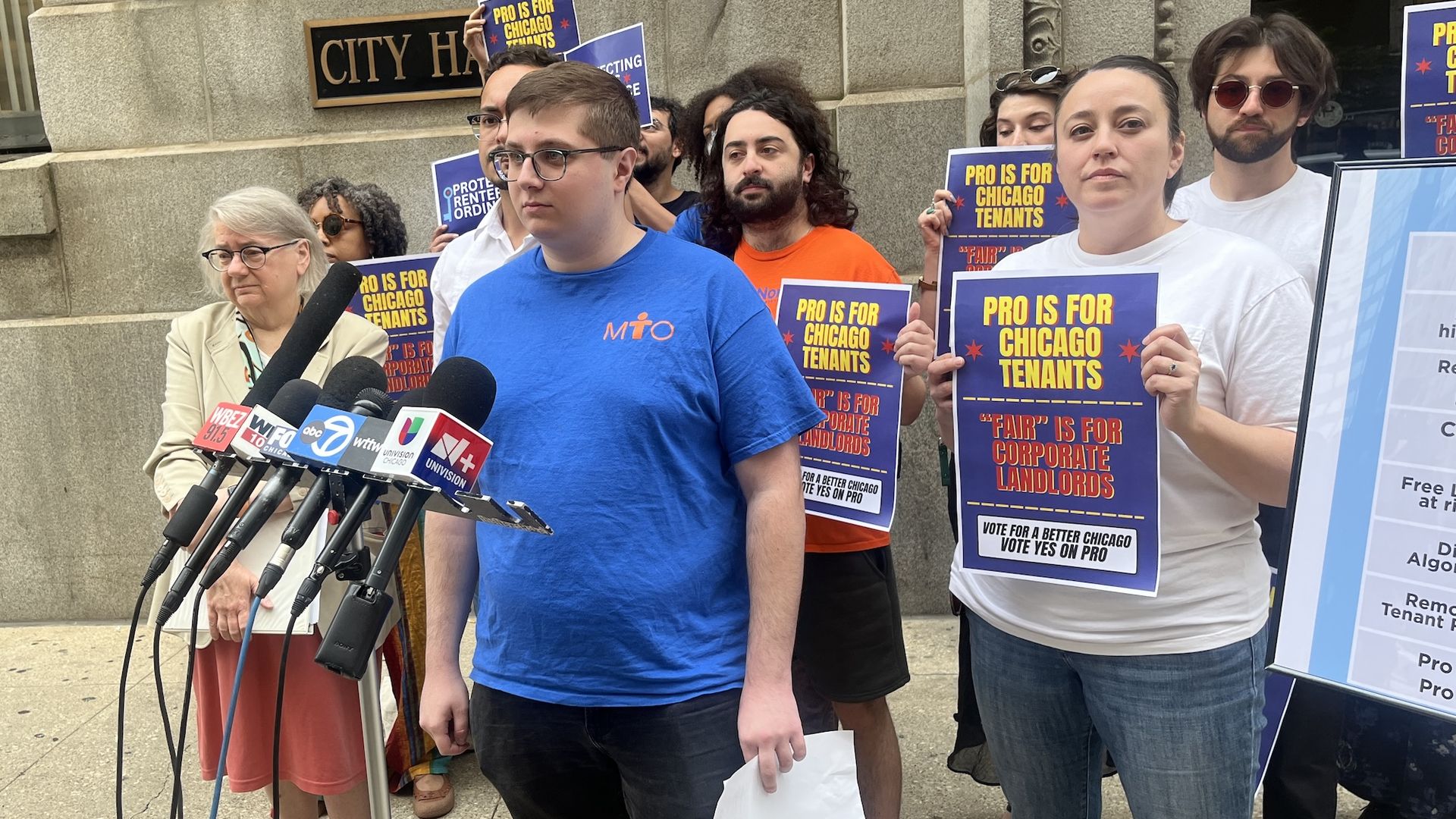 Group of protesters outside City Hall hold blue and yellow signs reading "PRO IS FOR CHICAGO TENANTS" while reporters with microphones stand nearby.