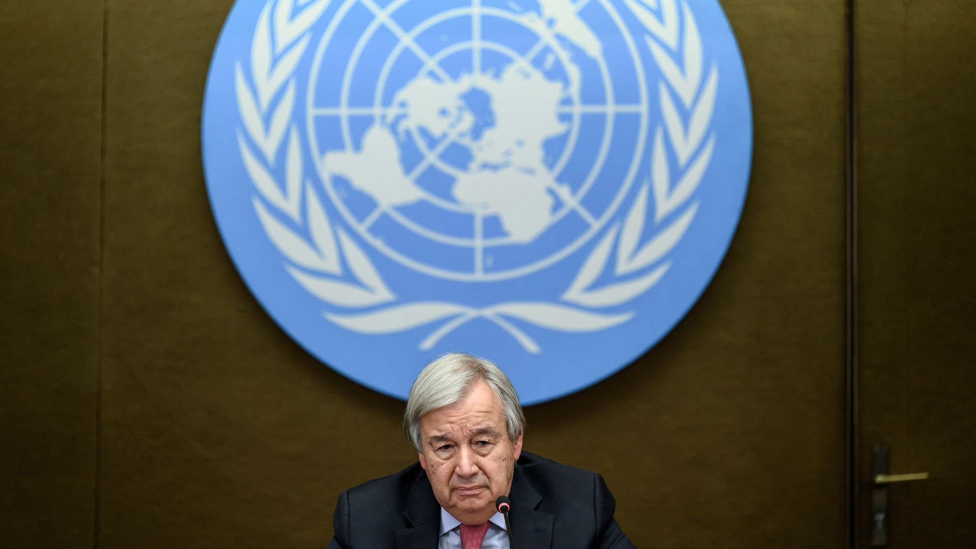 Photo of Antonio Guterres sitting with a giant plaque of the blue UN logo behind him