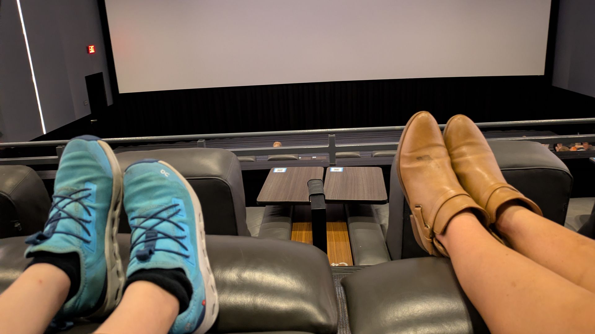 View from a movie theater: two people have their feet up in the seats. Left: teal sneakers with black socks; right: tan loafers. A large blank screen and dark curtains loom at the front.