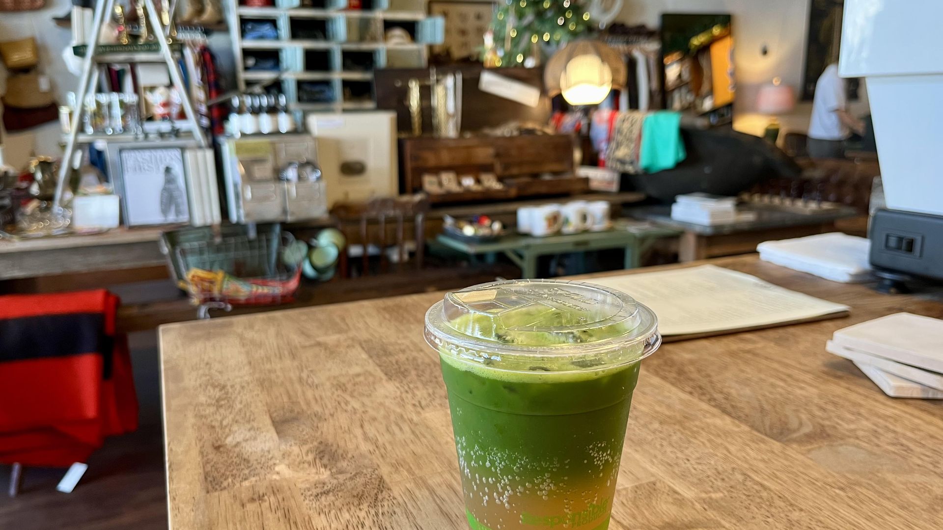 An iced green matcha layered on top of amber-colored apple cider in a clear plastic cup with a lid sits on a wooden table inside a cozy shop with shelves, clothes, and a small lit Christmas tree in the background. This is Wildcraft General on San Antonio's East Side.