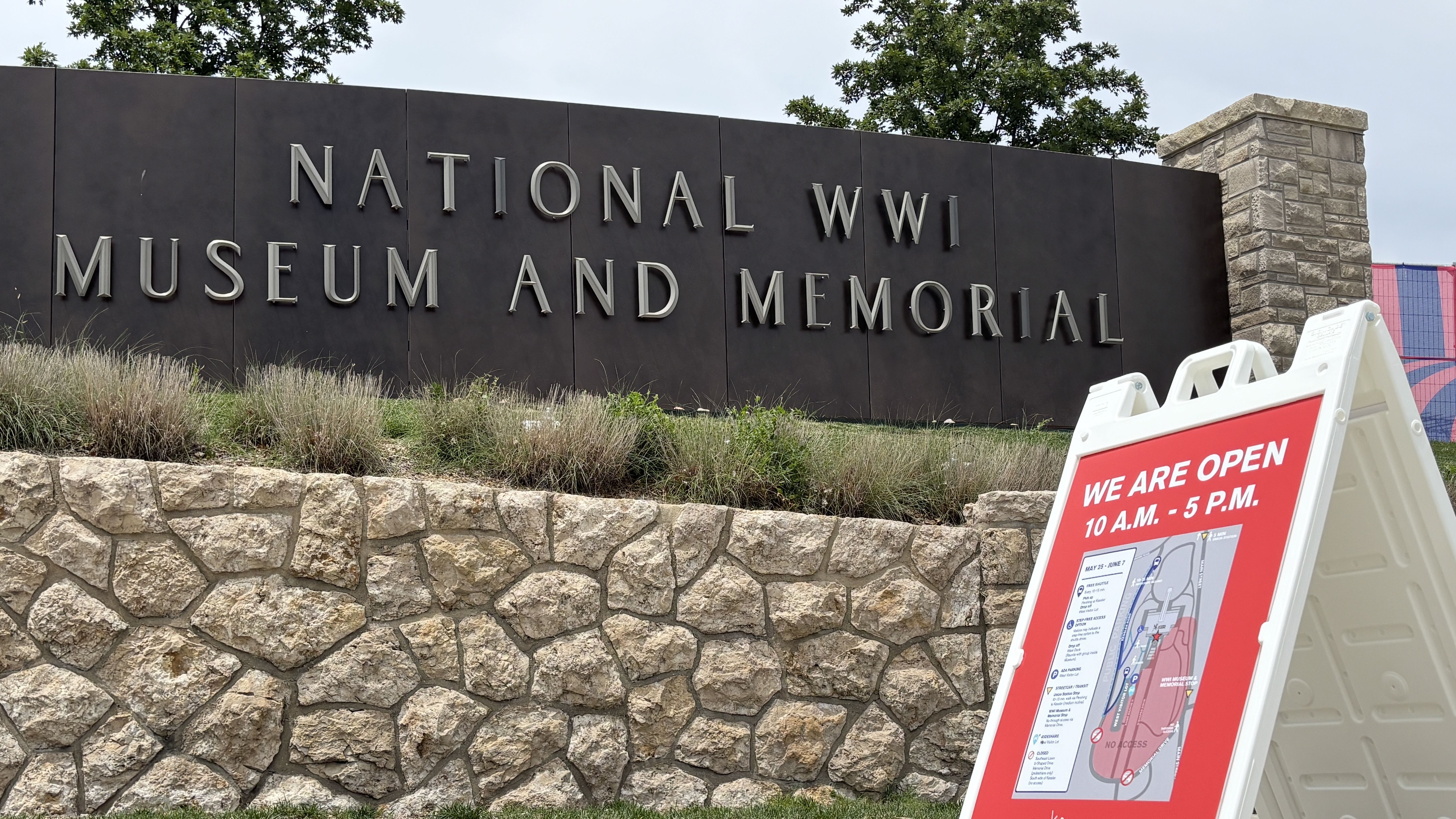Exterior view of the National WWII Museum and Memorial sign on dark panels with large silver letters, stone base, grasses and trees; a red sign in the foreground reads "WE ARE OPEN."