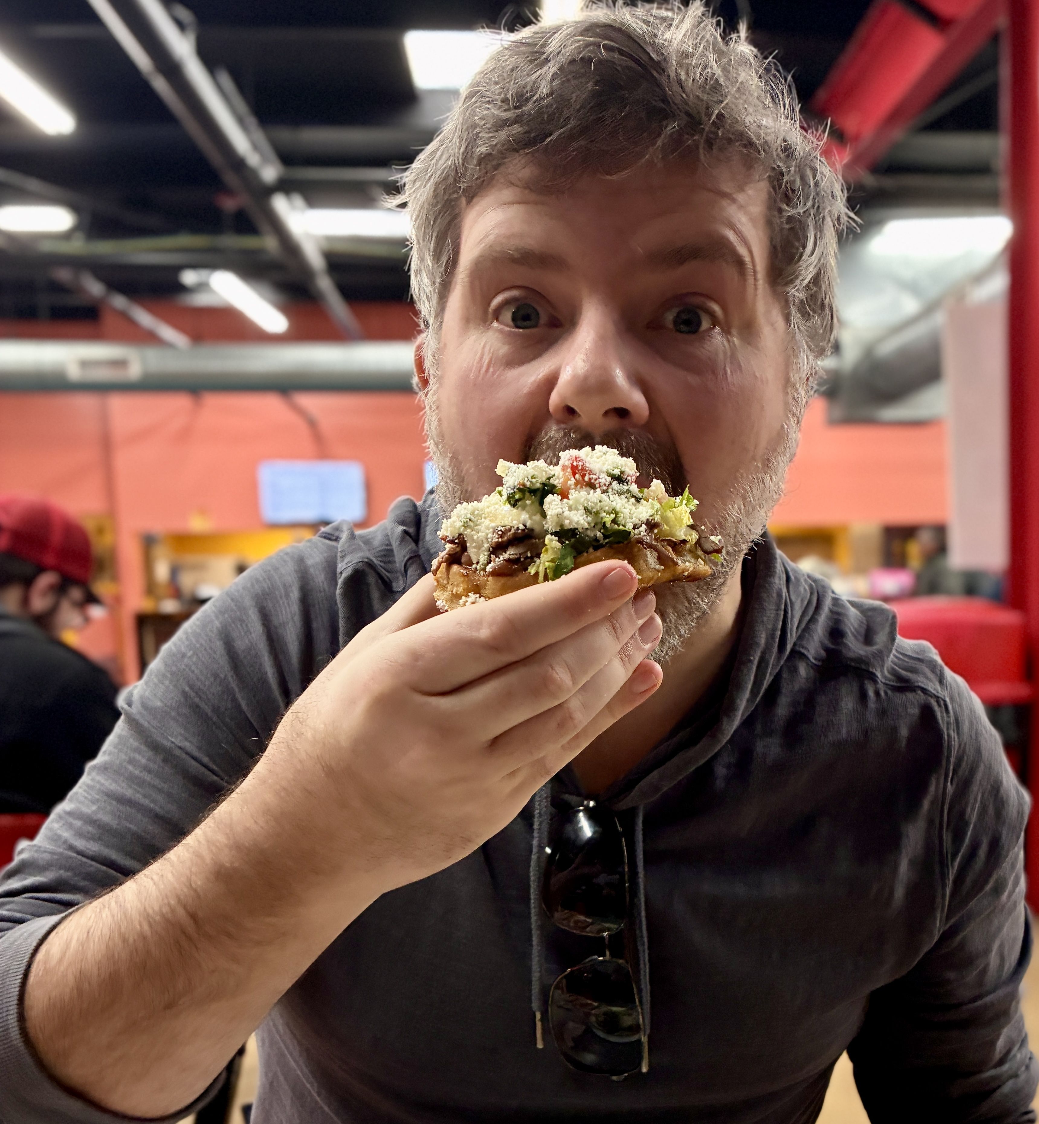 Close-up of Casey Weldon, a bearded man with gray hair taking a big bite from a loaded sope; wide-eyed expression, indoors at a casual restaurant with orange walls.