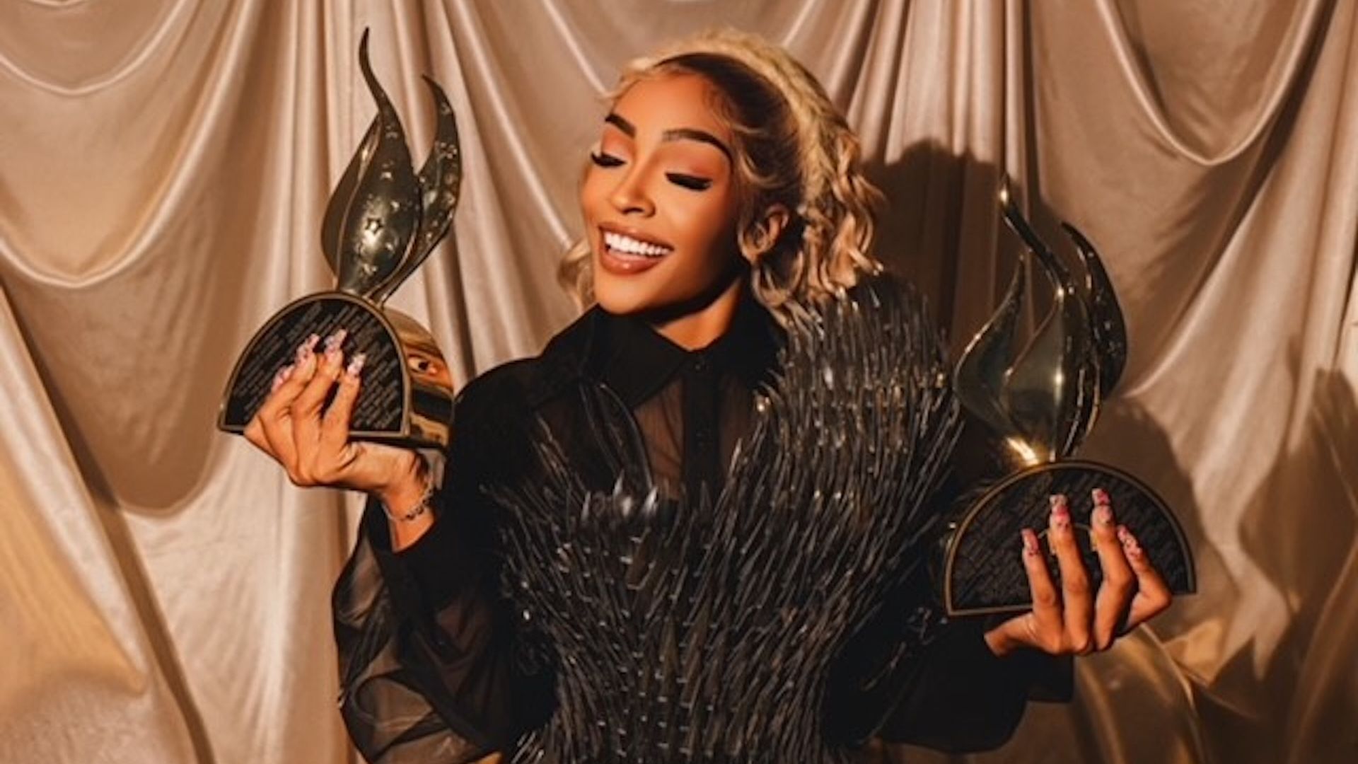 Smiling woman with long blonde hair in black dress holding two shiny trophies with a flowing beige curtain background.