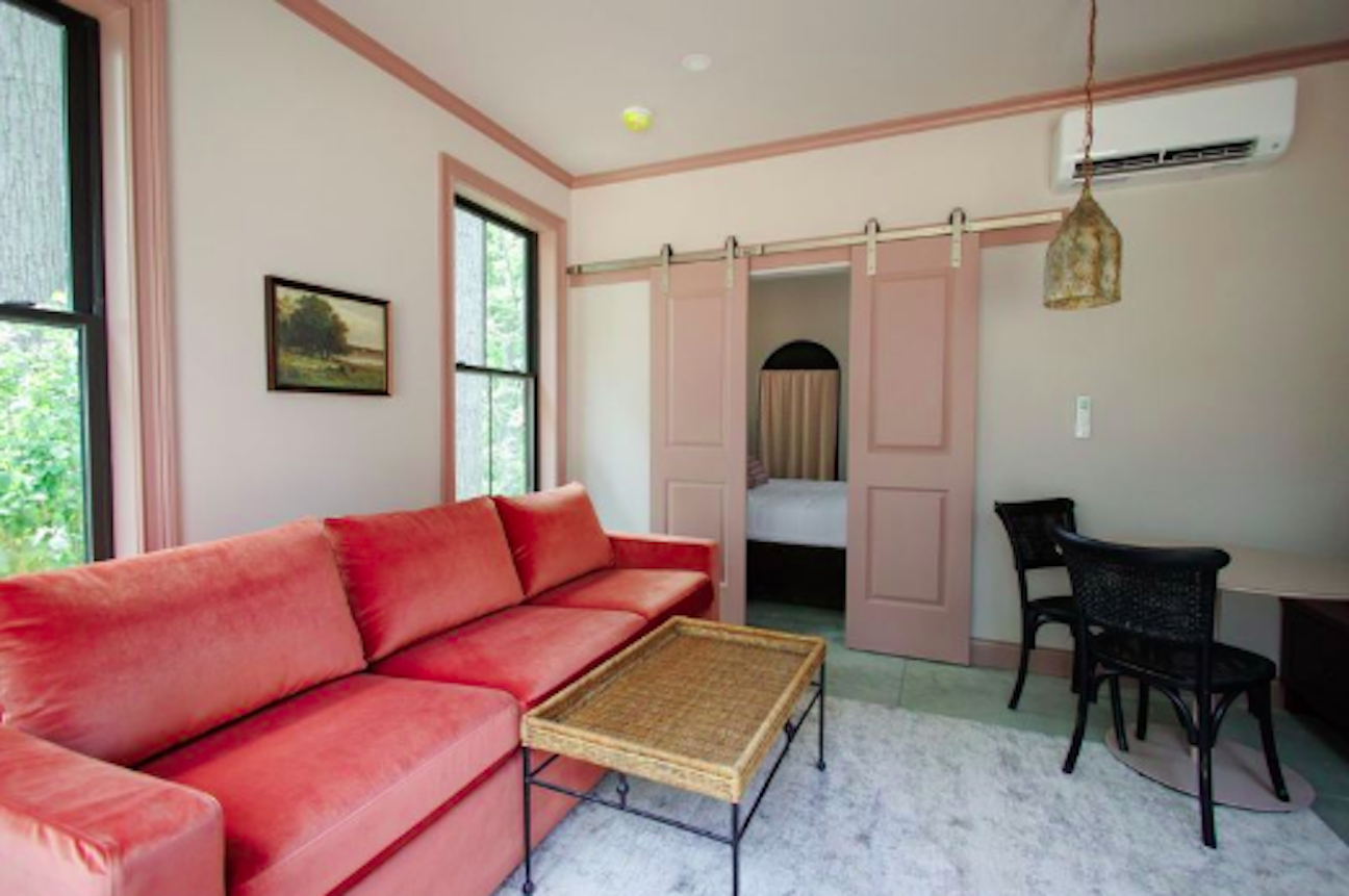 Cozy living room with pink walls and trim, coral sofa, woven coffee table, sliding pink barn doors to a bedroom, large windows showing greenery, a desk with black chairs, and a hanging lantern.