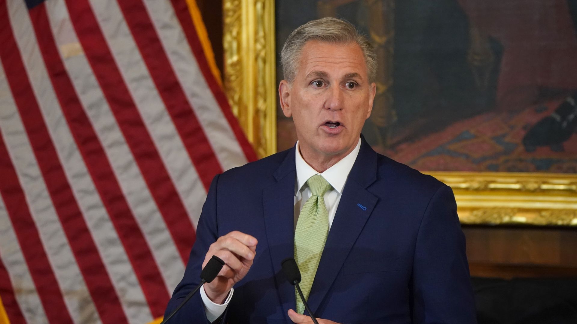 House Speaker Kevin McCarthy, wearing a blue suit, white shirt and green tie, speaks in front of an American flag and a large portrait.