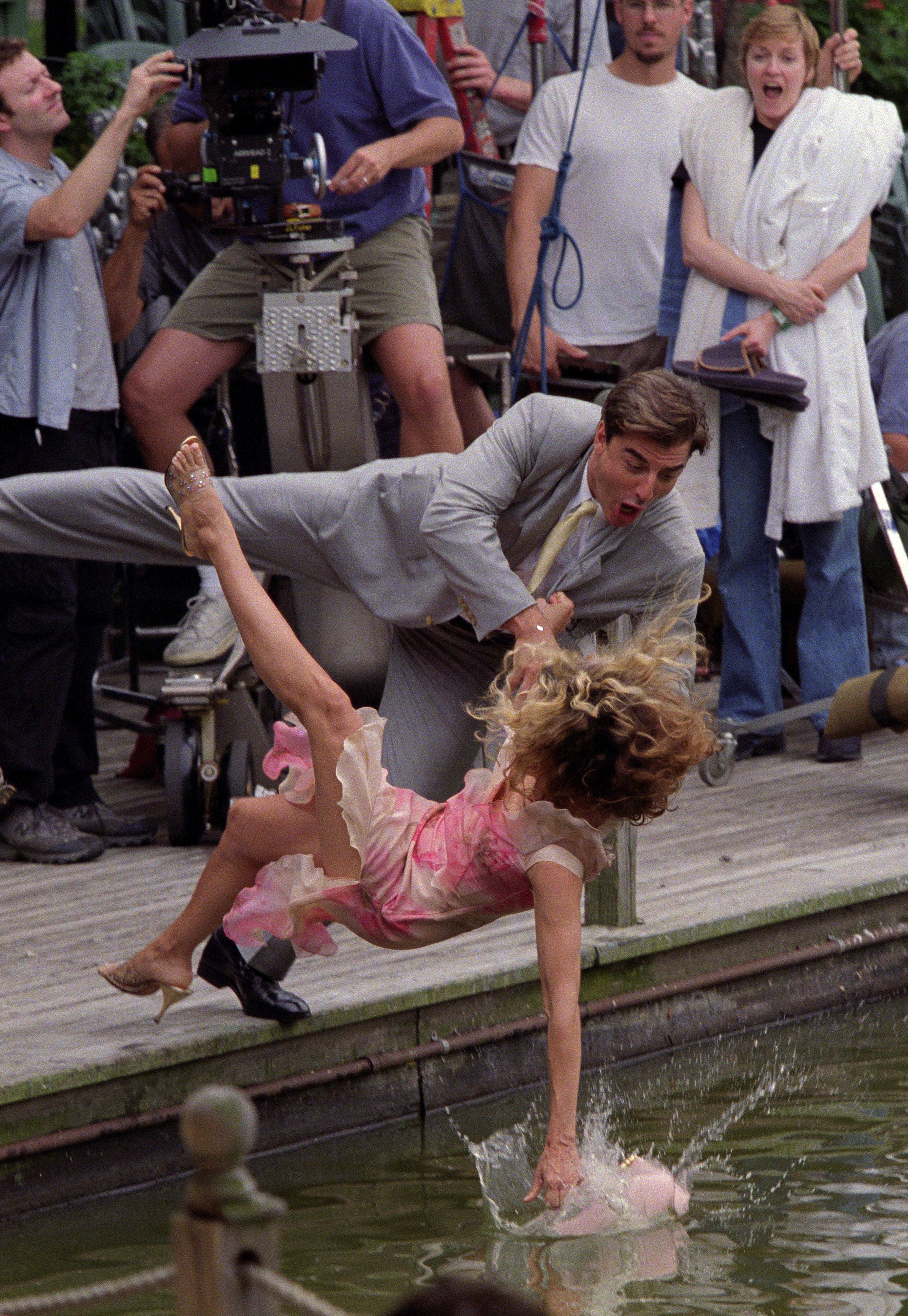 Scene being filmed with a man in a gray suit pulling a woman in a pink dress falling into water, while crew and onlookers watch in the background.