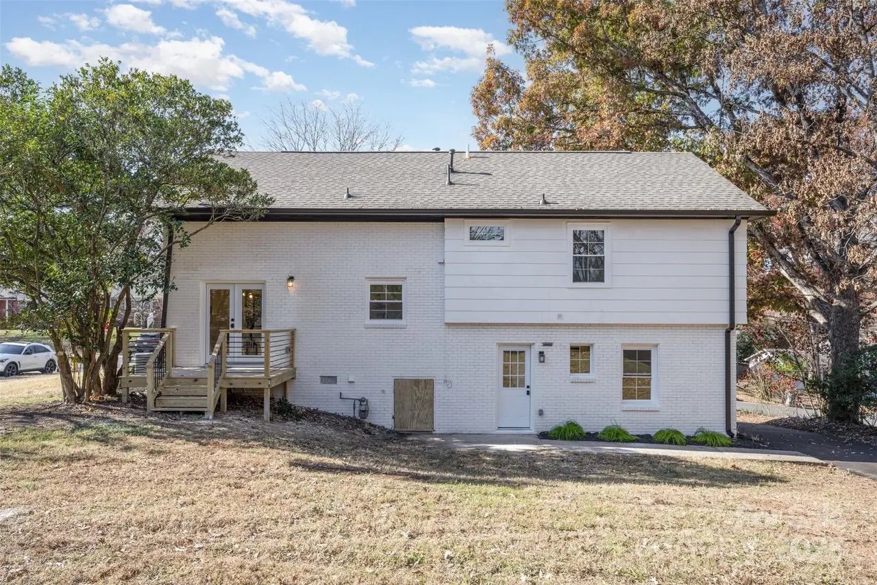 White two-story house with gray shingled roof, small wooden porch, and surrounding trees with autumn and green leaves under a partly cloudy blue sky.