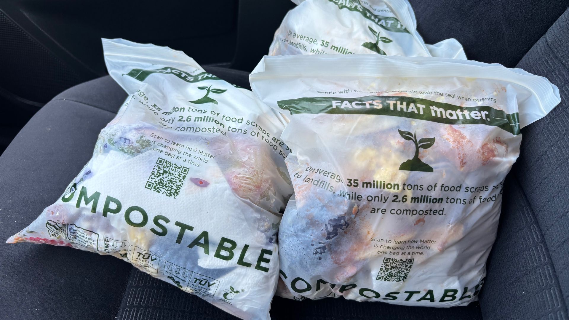 Inside a car, several crumpled white compostable bags with green text and a plant logo lie on the dark seat; labels read "FACTS THAT MATTER" and they contain food scraps.