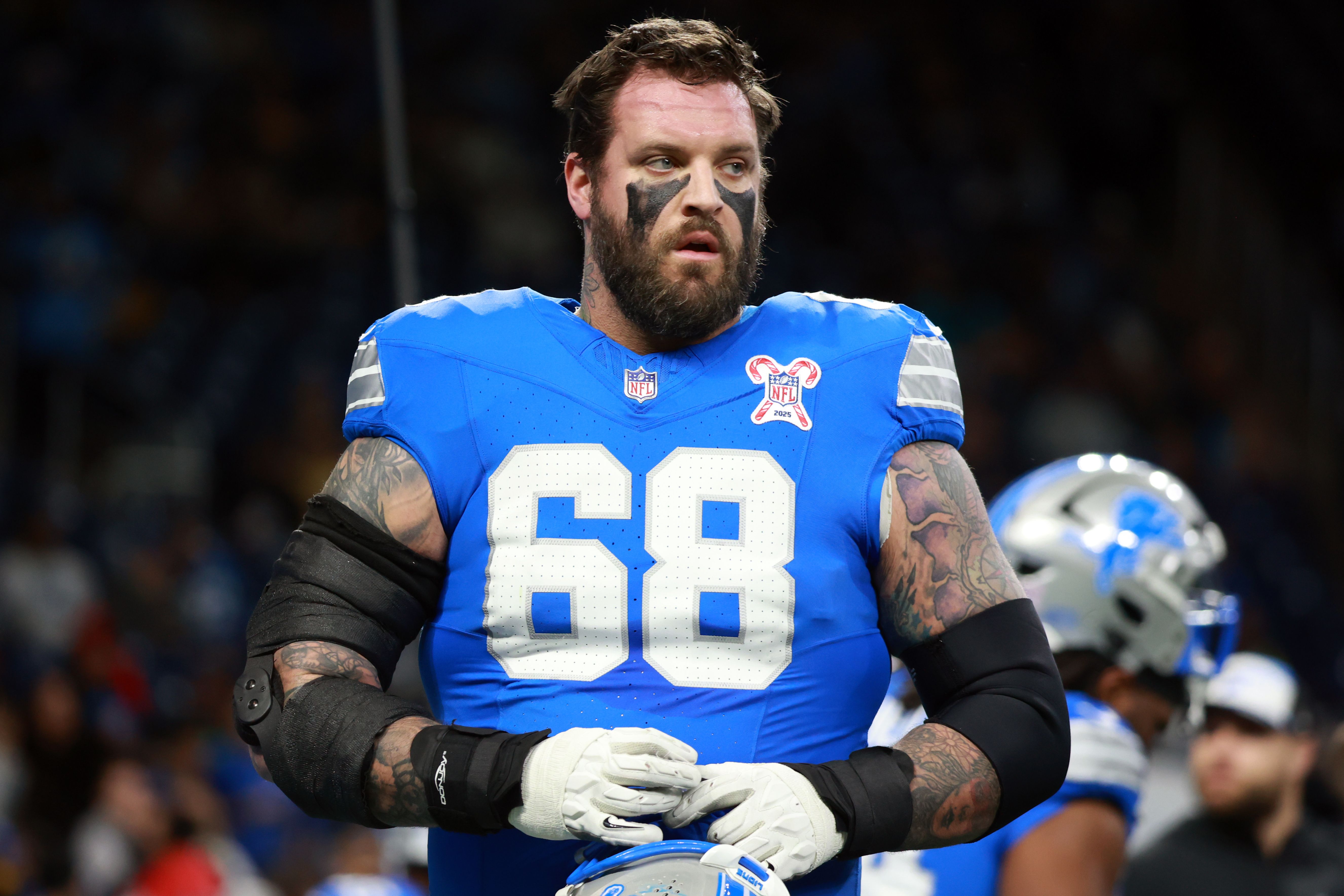 Bearded American football player in a blue jersey with number 68, black eye paint, and tattoos on arms, holding a helmet during a game with blurred players in the background.
