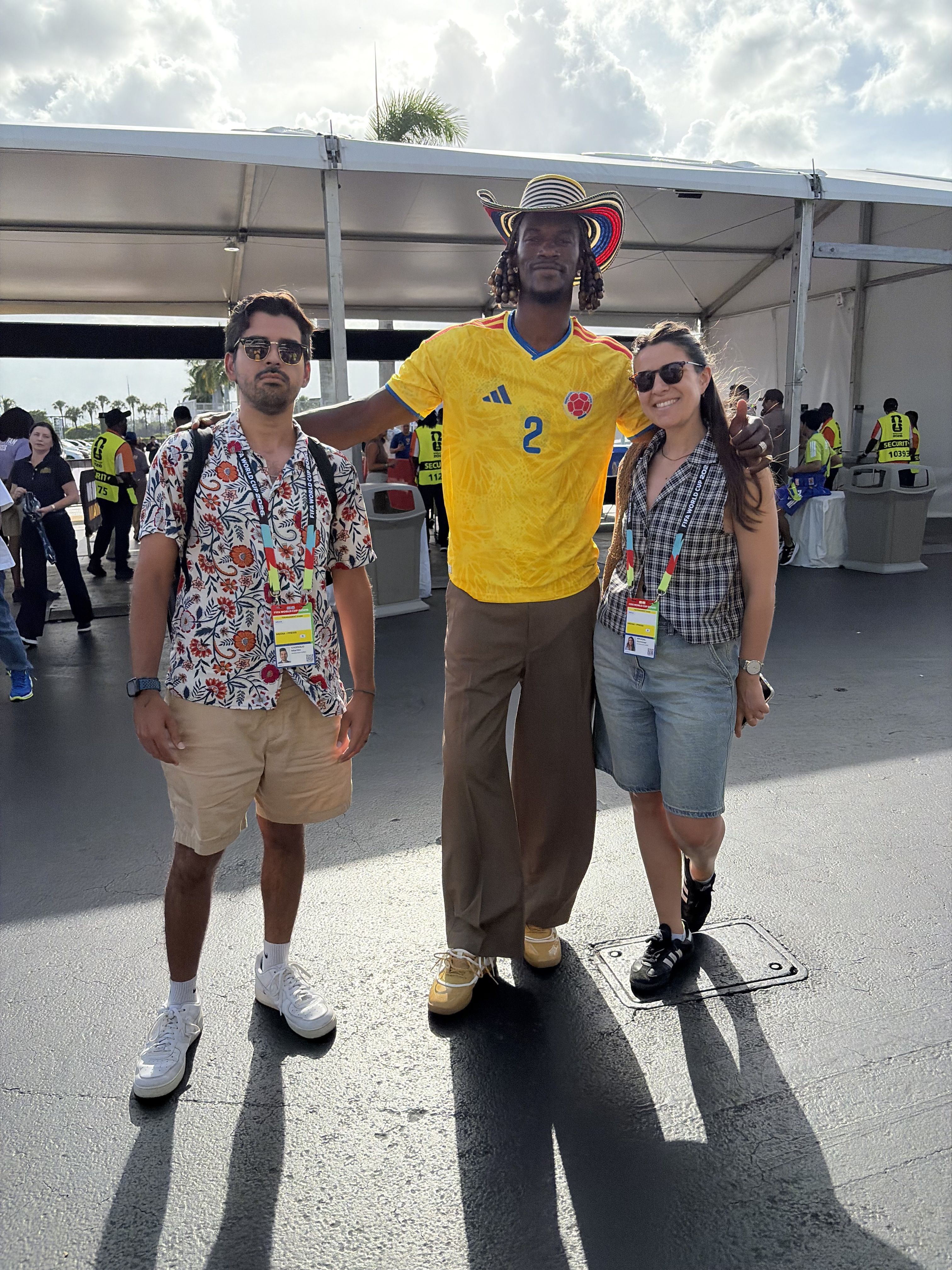 Three people pose under a white event tent: a floral shirt man, a tall man in a yellow Colombia jersey with a colorful sombrero, and a plaid-top woman in denim shorts, all wearing lanyards.