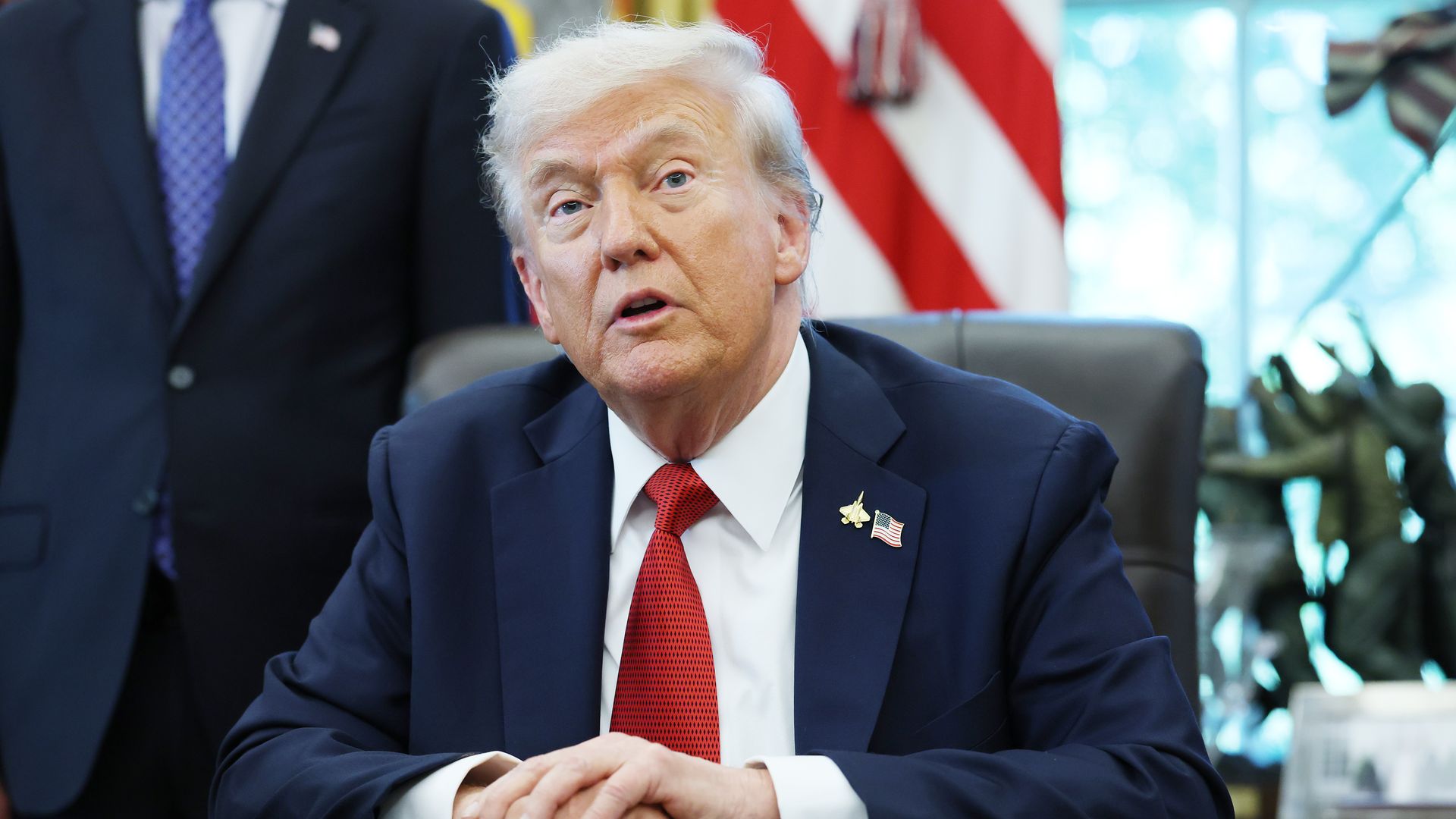 President Trump, wearing a navy jacket with a US flag pin and a gold plane pin, white shirt and red tie, speaks in the Oval Office of the White House from a black chair in front of a US flag and the body of an unidentified man in a navy suit, white shirt and blue and white-check tie.