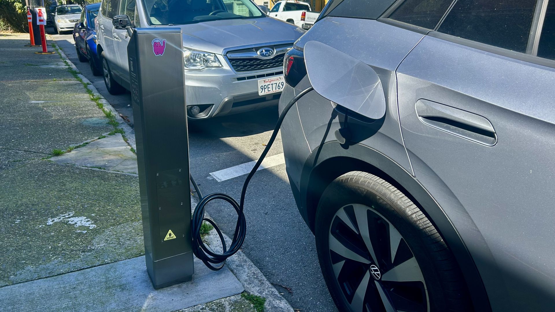 City street with a tall black EV charging pedestal on the sidewalk; a silver SUV on the right is plugged in, with coiled cables, while colorful row houses line the background.