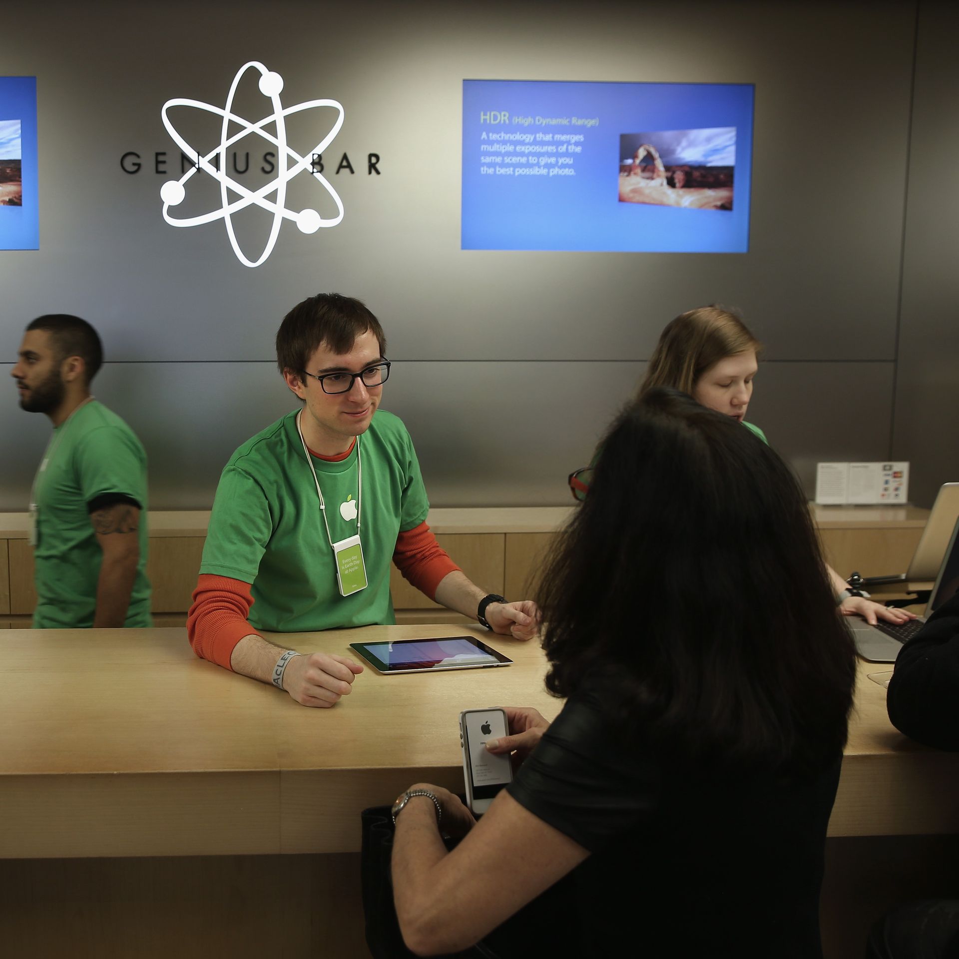 A man in a green shirt, layered over a red long-sleeved shirt, leans over a counter in front of a sign that says "Genius Bar"