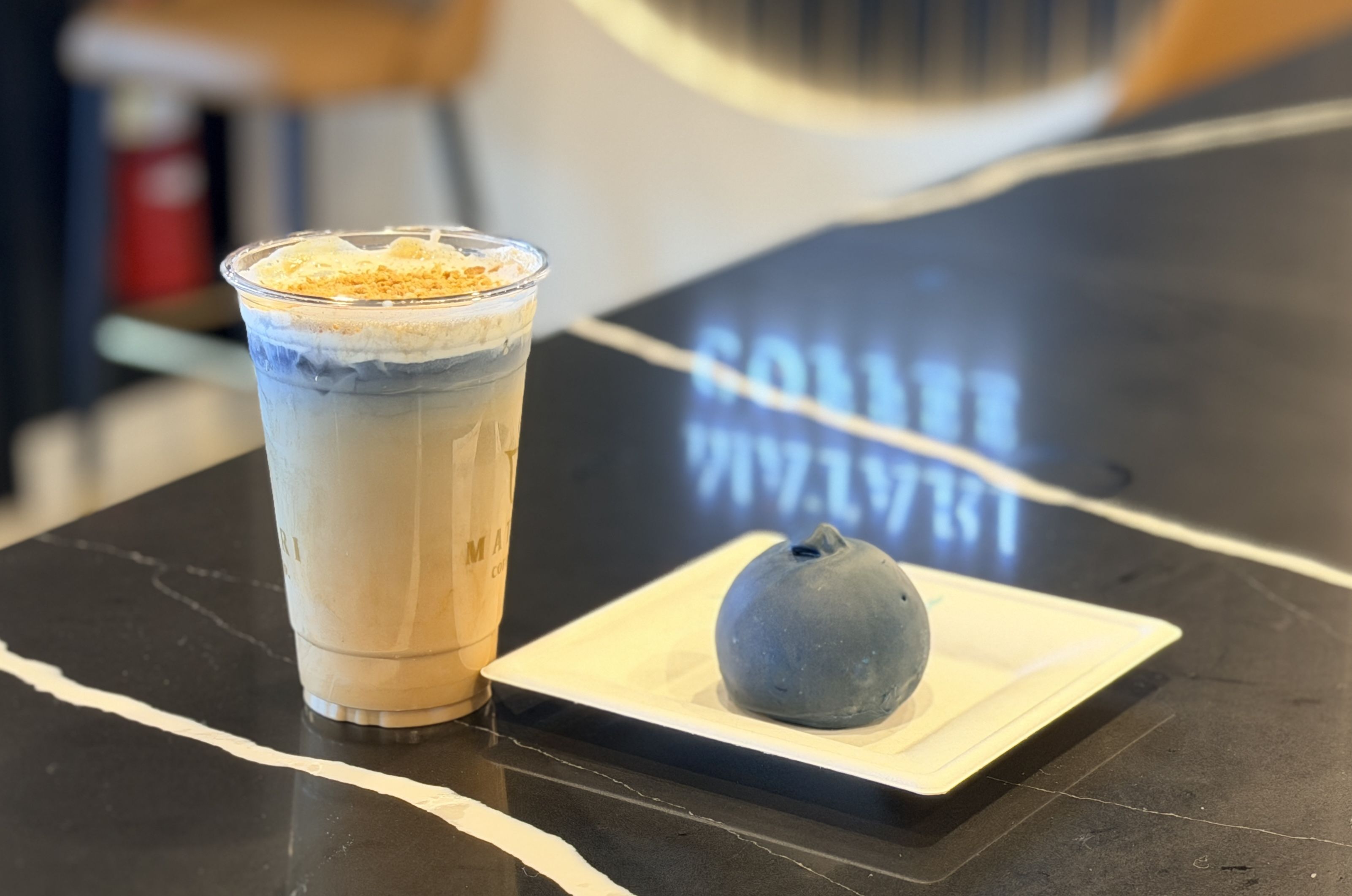 Tall plastic cup of blue iced drink with whipped cream and orange crumble sits on a dark marble table; in front are two blue spheres on a white tray, blurred in the background.