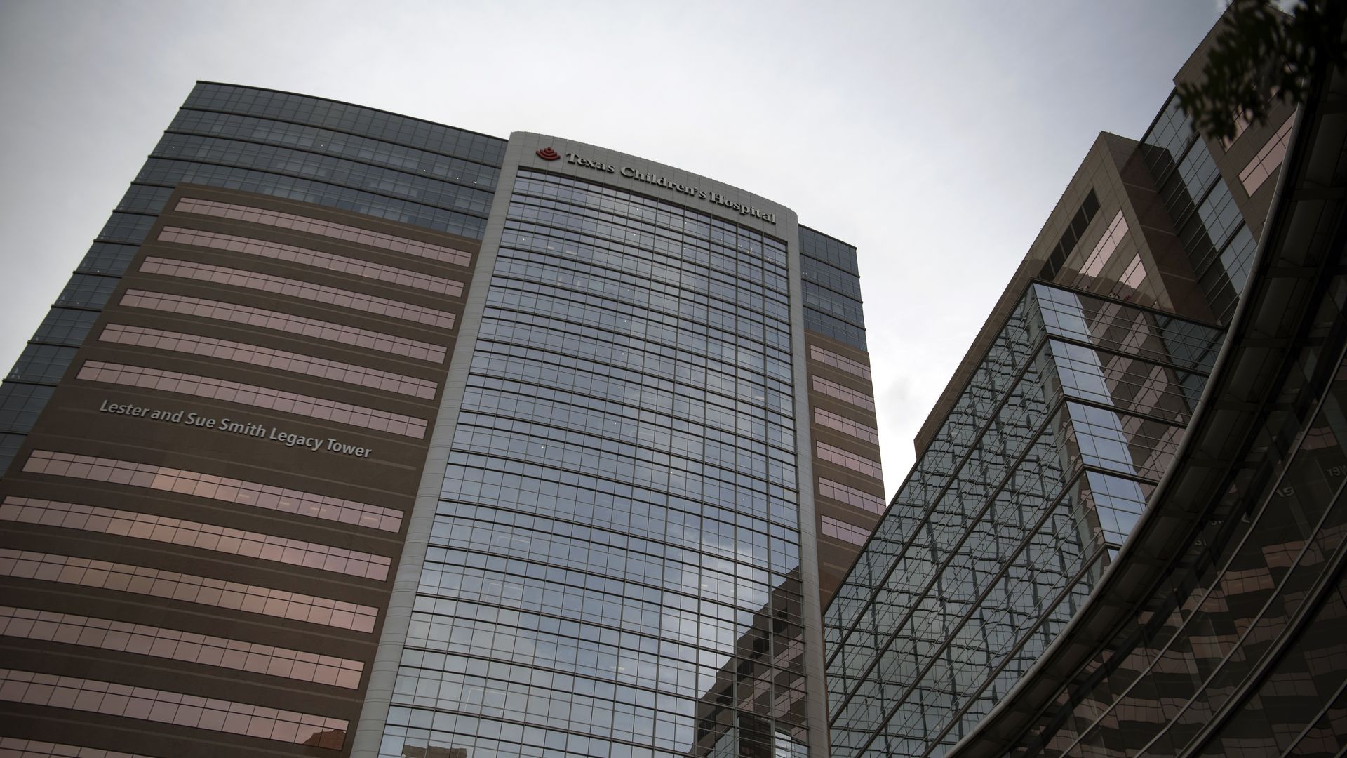 Glass-front hospital campus with a tall central tower labeled "Texas Children's Hospital" and a red logo; lower tower reads "Lester and Sue Smith Legacy Tower" against a pale sky.