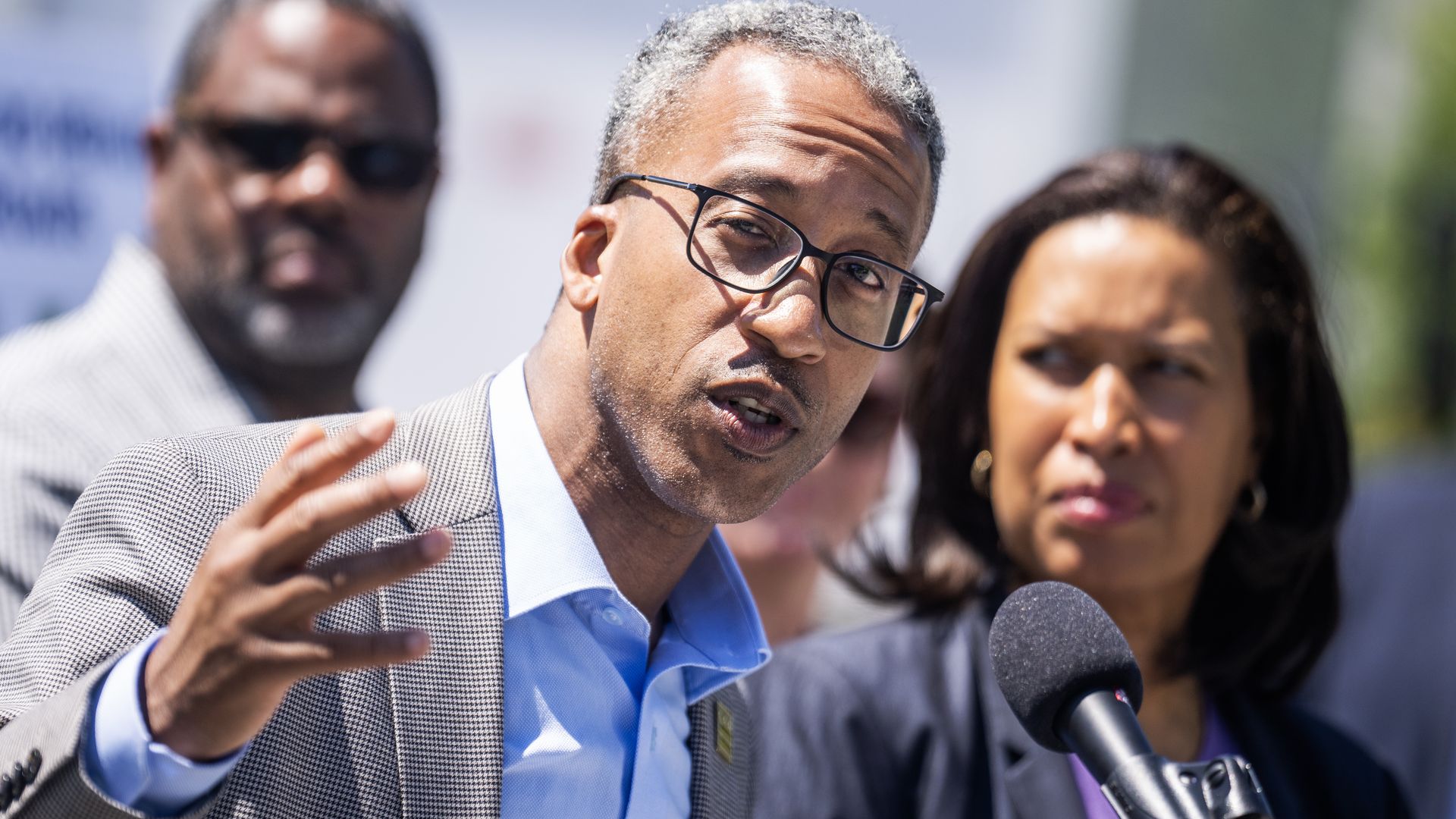 Kenyan McDuffie talks in front of a podium, with Mayor Muriel Bowser next to him in the background