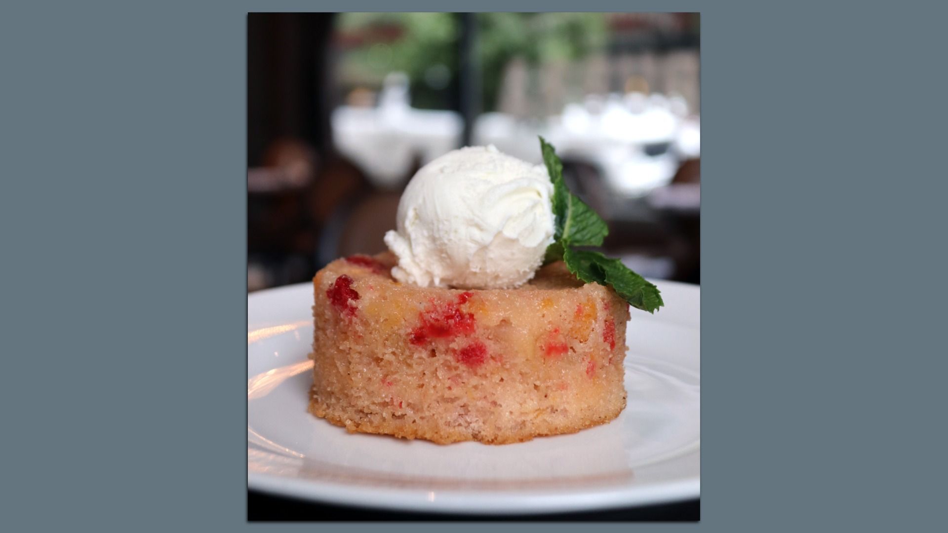 A square slice of sponge cake studded with red berries, topped with a scoop of vanilla ice cream and a mint leaf, served on a white plate with a softly blurred restaurant backdrop.
