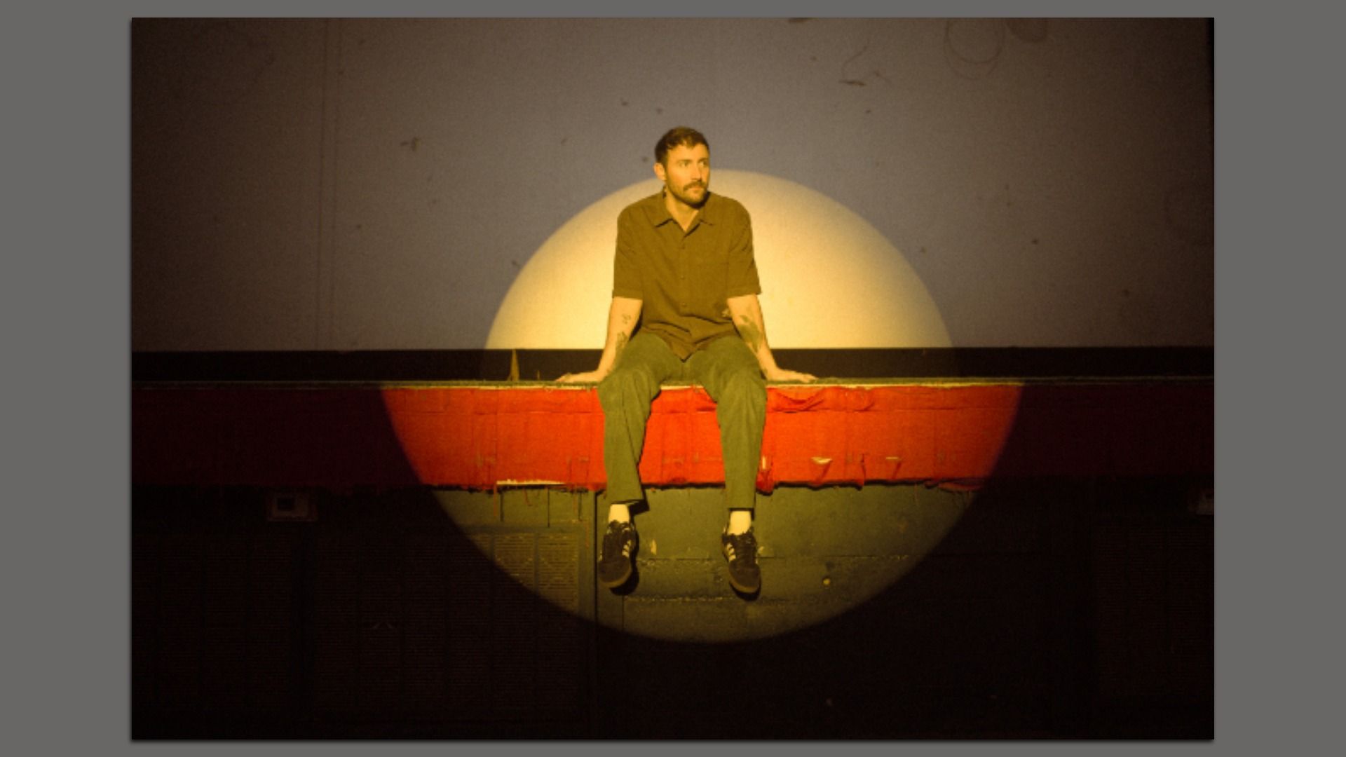 A man with a beard sits on a red ledge under a spotlight against a dark background, wearing a brown shirt, green pants, and black sneakers.