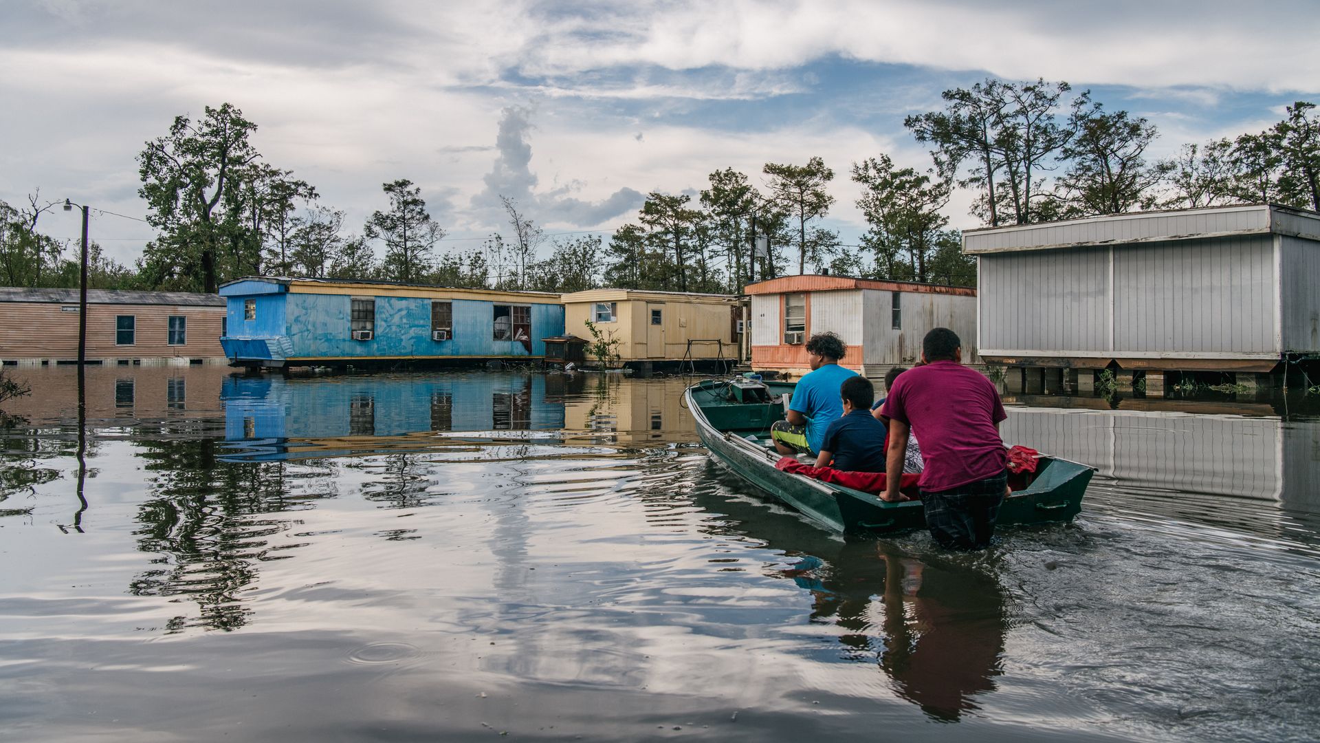 A family of four, with their backs to the camera, motors along in a small boat through a flooded neighborhood. Mobile homes and a cloudy sky can be seen in the background.