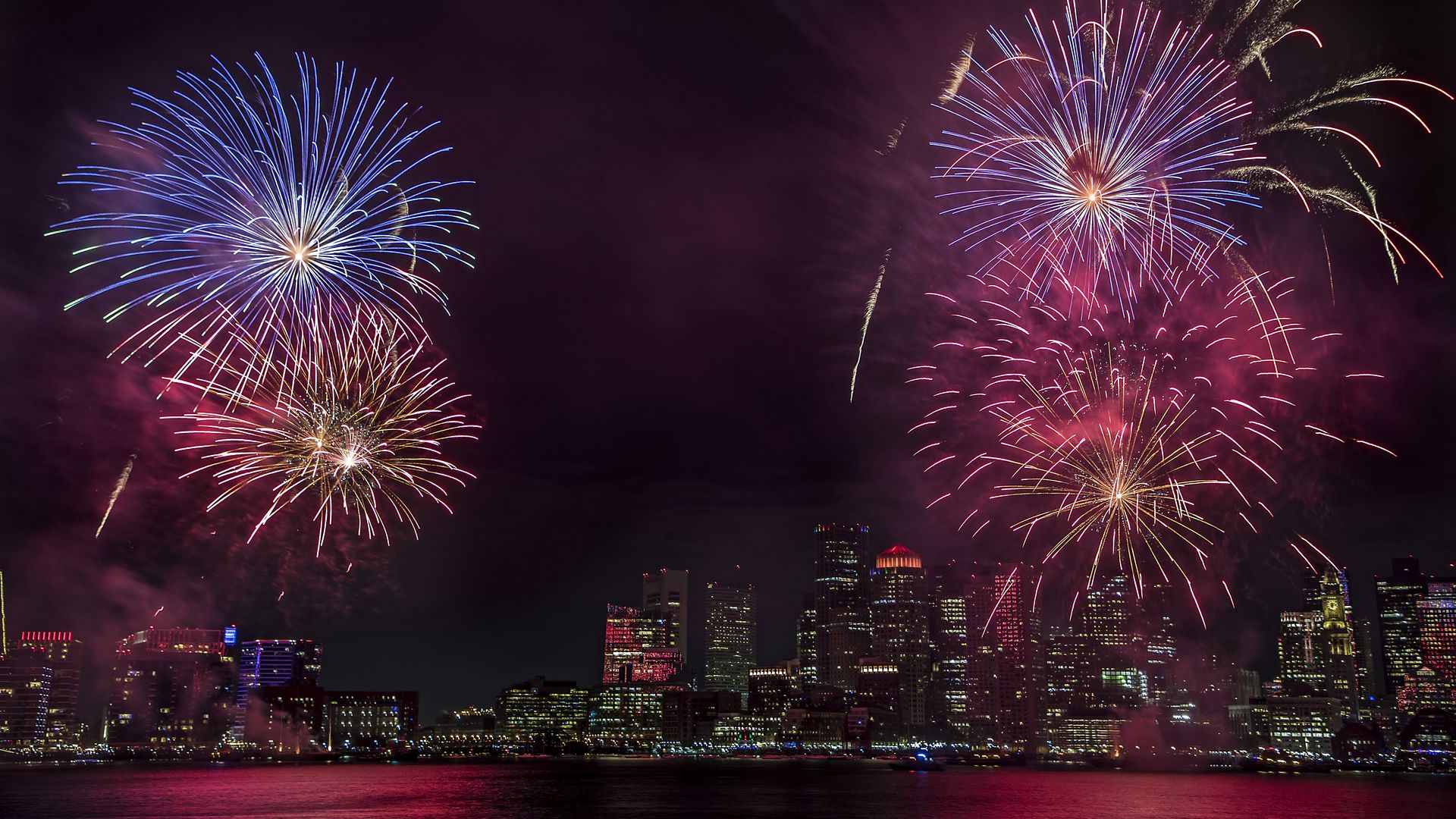 Colorful blue and pink fireworks exploding over a city skyline at night, reflecting off the water below with buildings lit up in the background.