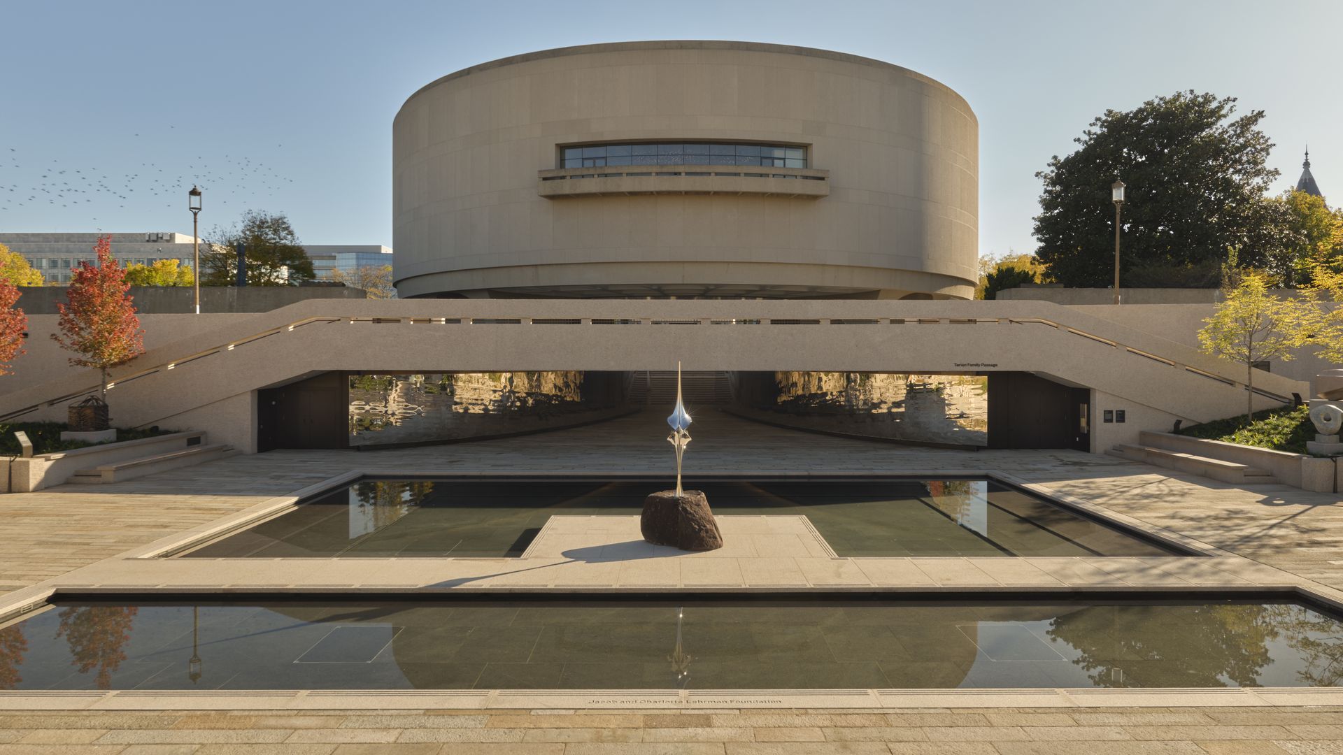 Wide view of a circular beige museum with a curved façade and a long central balcony window. In front, a reflecting pool with a sculpture, staircases, trees, and a clear blue sky.