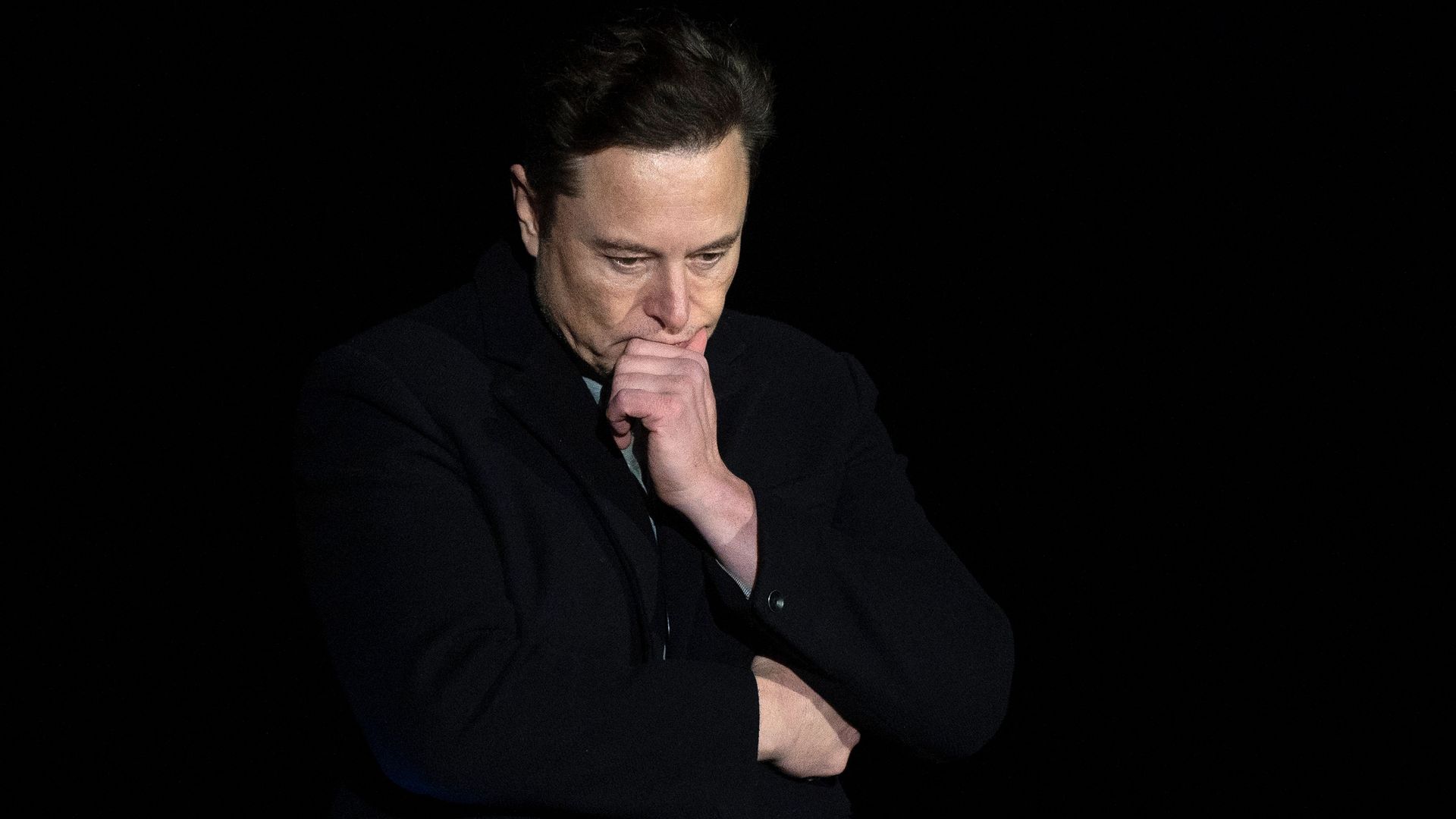 Photo of Elon Musk with his hand on his chin against a black background