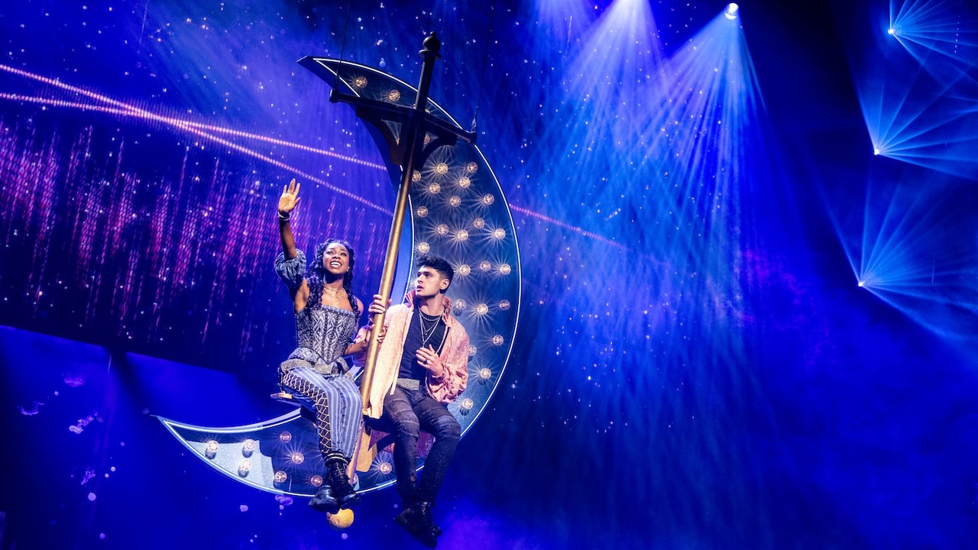 Two performers in costume sit on a glowing crescent moon prop suspended on a stage with blue starry lighting and spotlights creating a magical atmosphere.