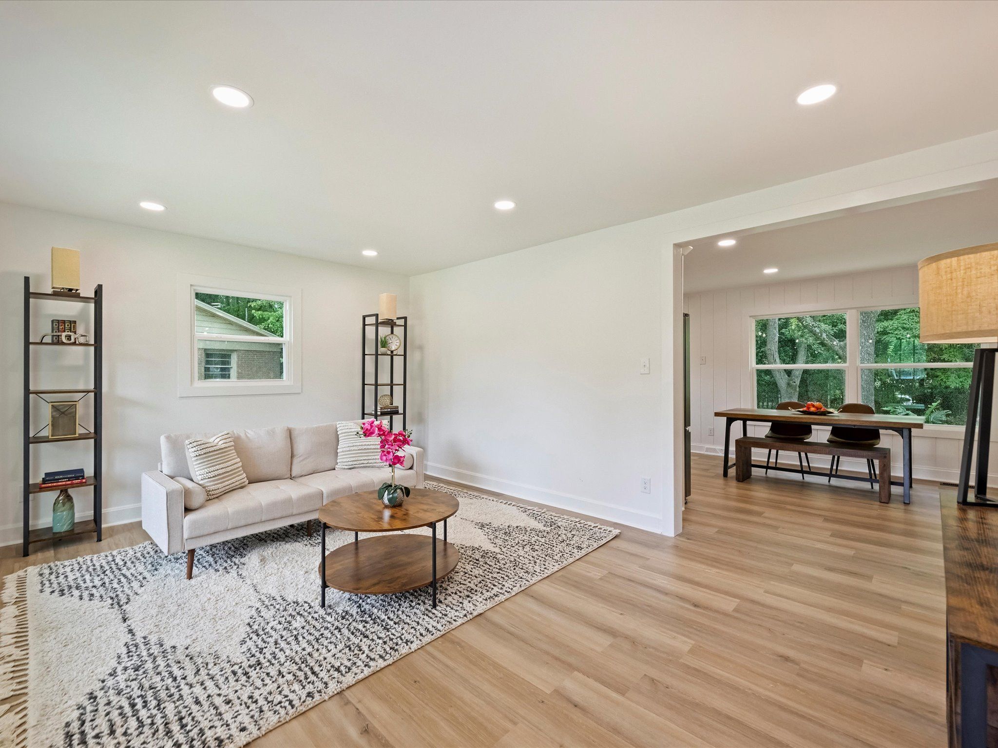 Bright living room with beige couch, patterned rug, round wooden coffee table holding pink flowers, black metal shelves with decor, adjacent dining area with wooden table and large window.