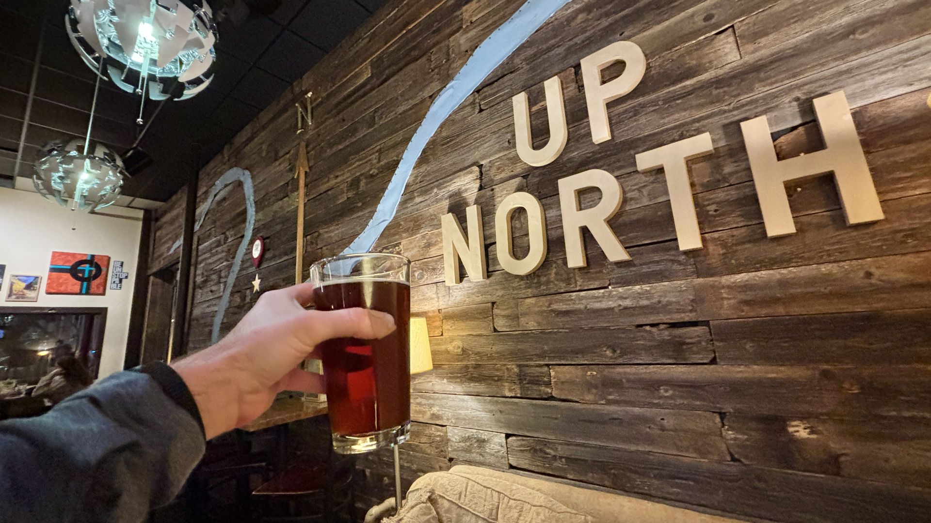 Hand holding a glass of dark beer inside a cozy bar with wooden wall art spelling "UP NORTH" and a blue painted river and compass arrow.
