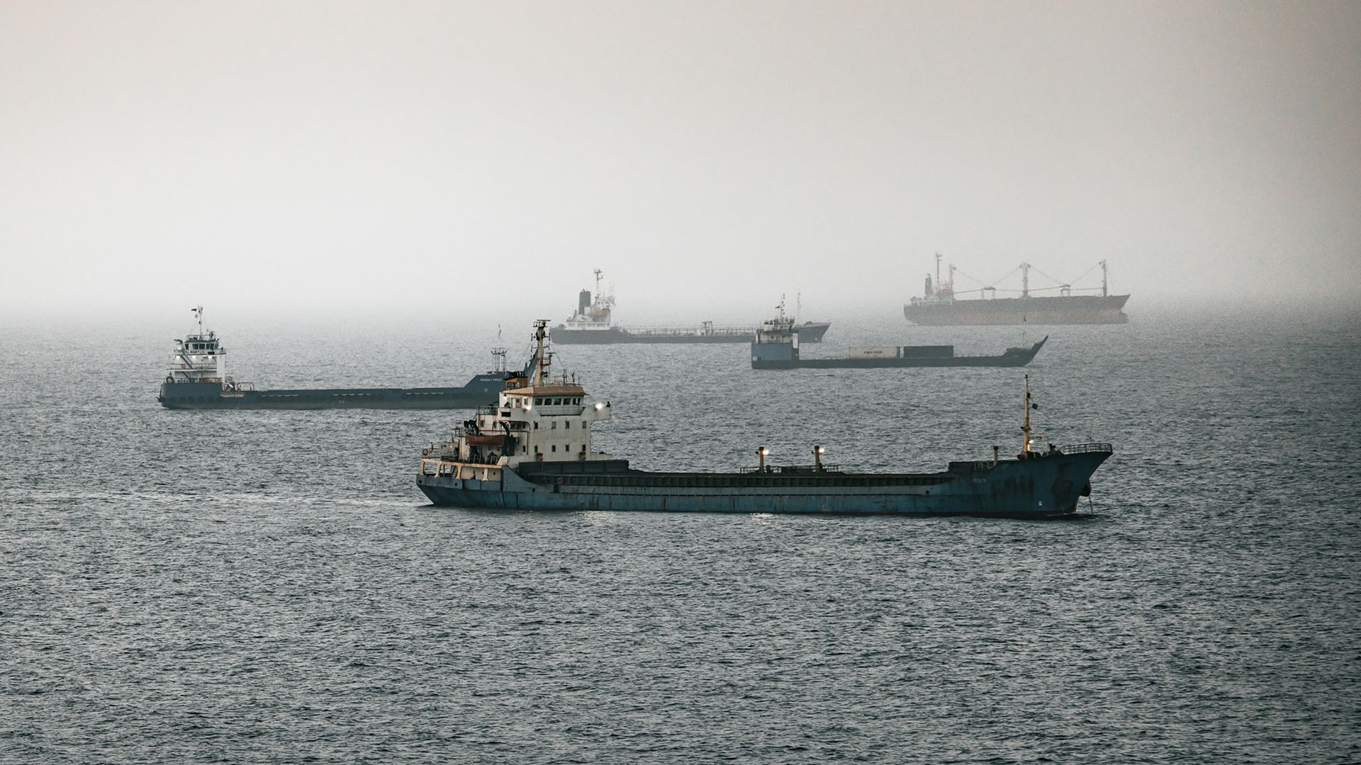 Calm sea with several cargo ships and barges, some in the foreground and others on the hazy horizon; distant ships fade into a pale gray sky.